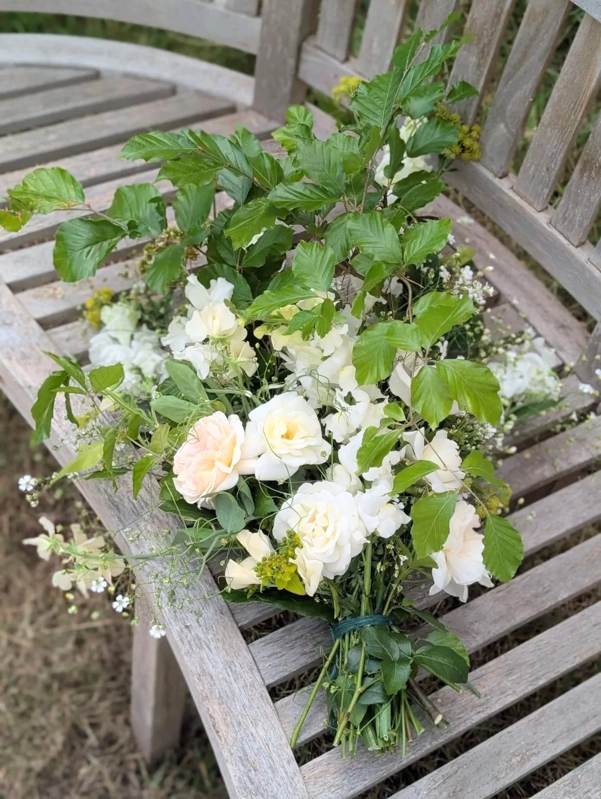Farewell spray arrangement of white and pale pink flowers with green leaves resting on a weathered wooden bench.