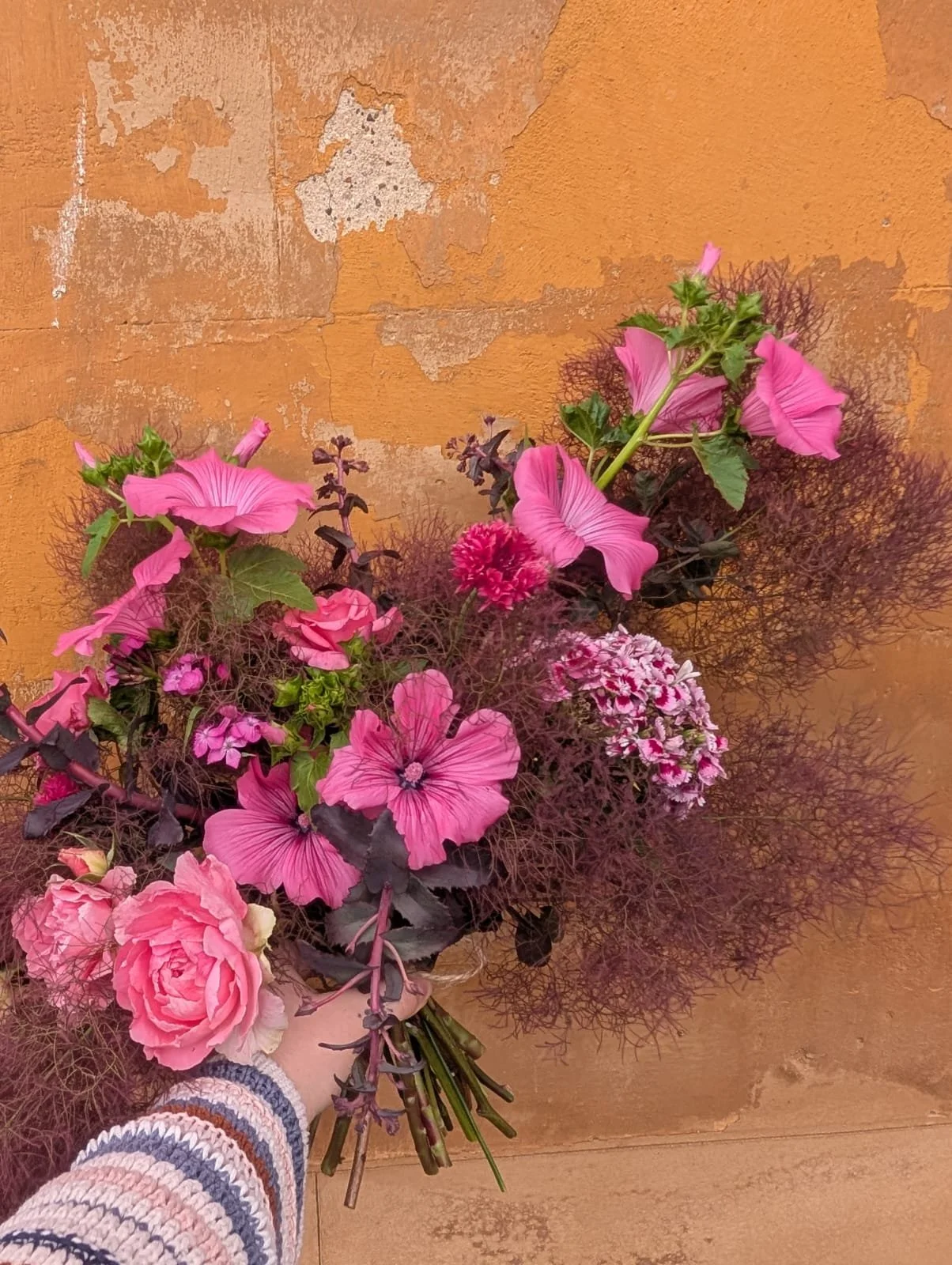 A person holding a mixed bouquet of pink and purple flowers, including petunias, roses, and other blooms, against a weathered orange wall.