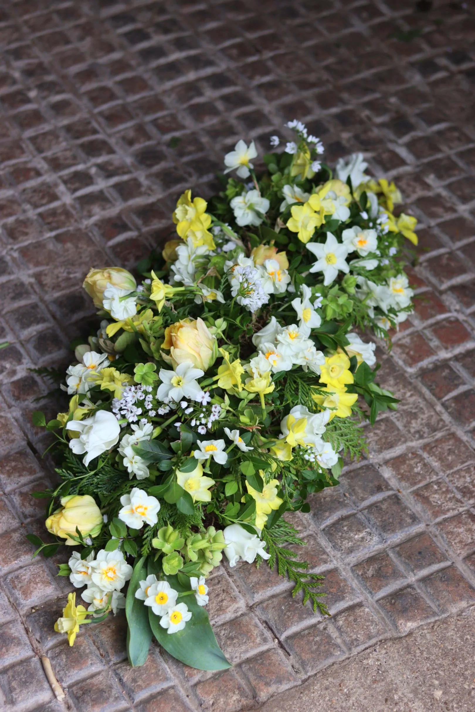 Farewell spray arrangement with white and yellow flowers on a brick floor.