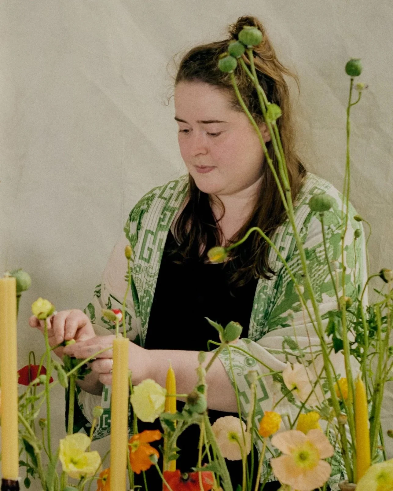 A woman with wavy brown hair styled in an updo, wearing a beige jacket with green geometric patterns, is sitting at a table with various colorful flowers and yellow birthday candles, focusing on arranging them.