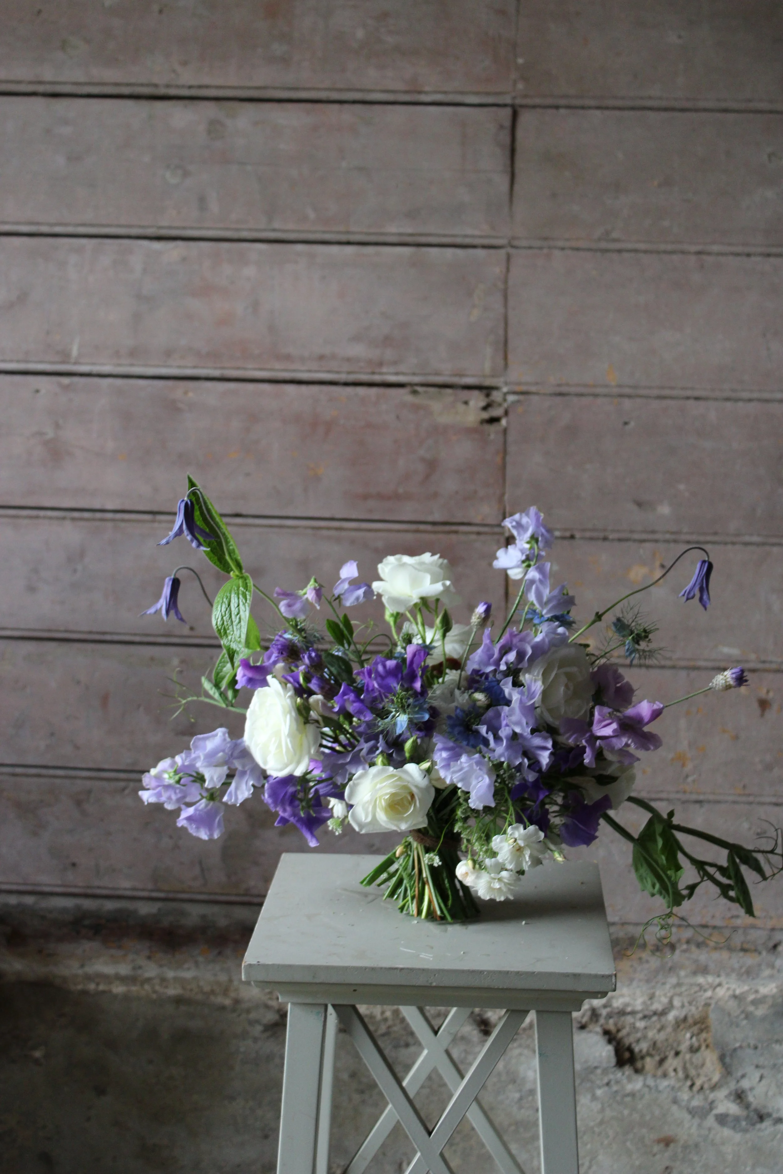 A bouquet of white and purple flowers arranged in a glass vase on a white table against a wooden wall background.