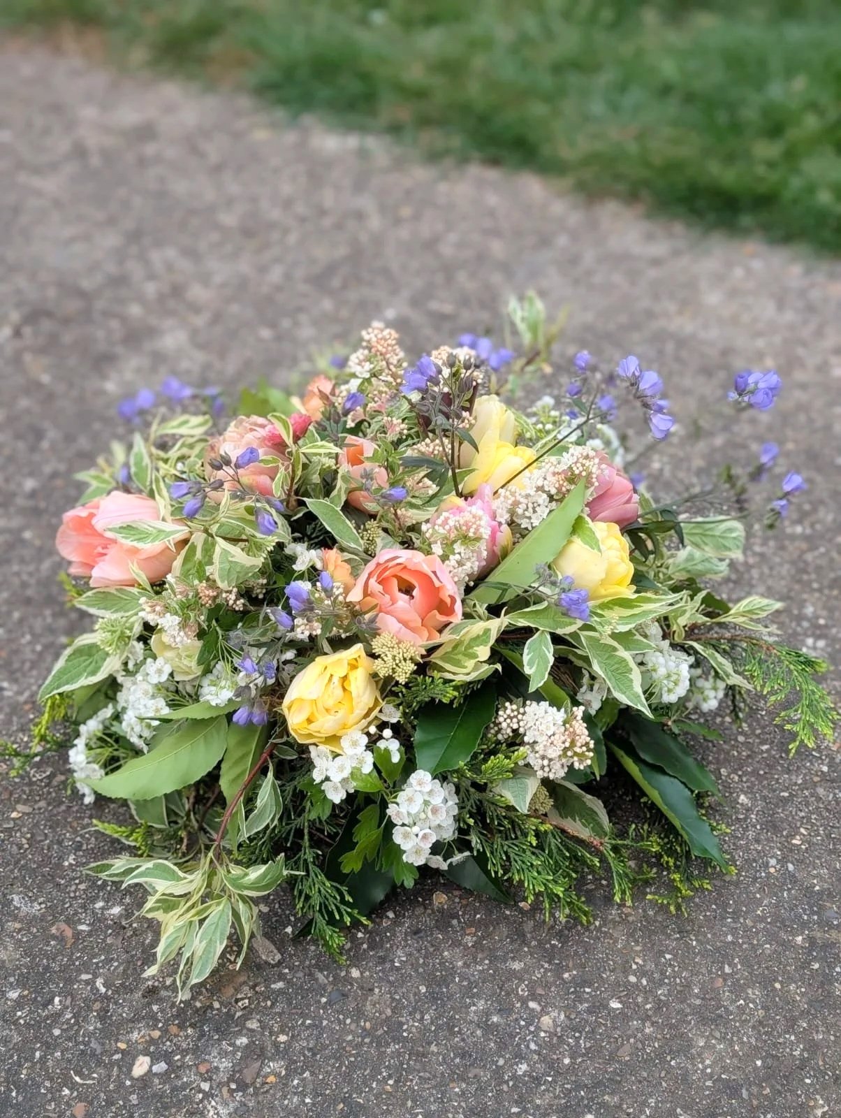 Farewell wreath arrangement with pink, yellow, and purple flowers, and green foliage, placed on a gray surface outdoors.