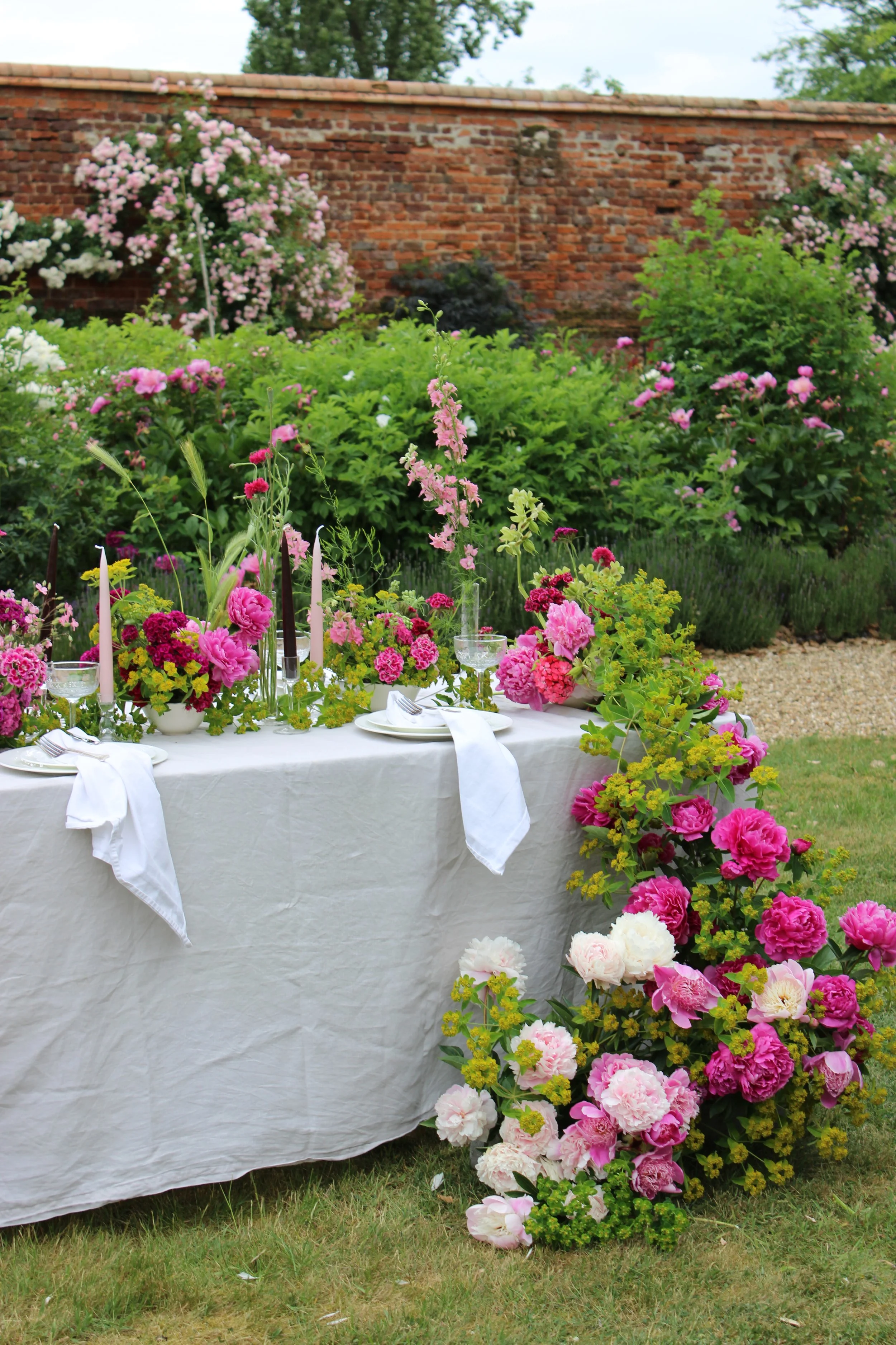 A table decorated with pink and white flowers, set outdoors on grass with a white tablecloth, and accented with pink candles, in front of a brick wall with green bushes and trees.