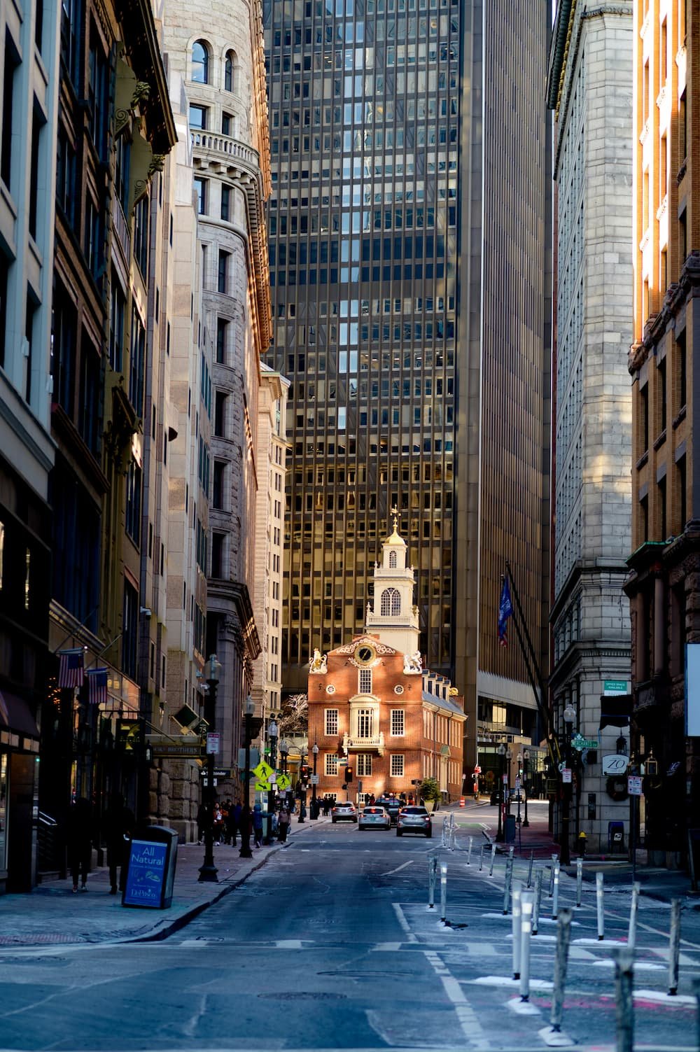 A city street with tall buildings on both sides and a historic brick building with a clock tower in the center.