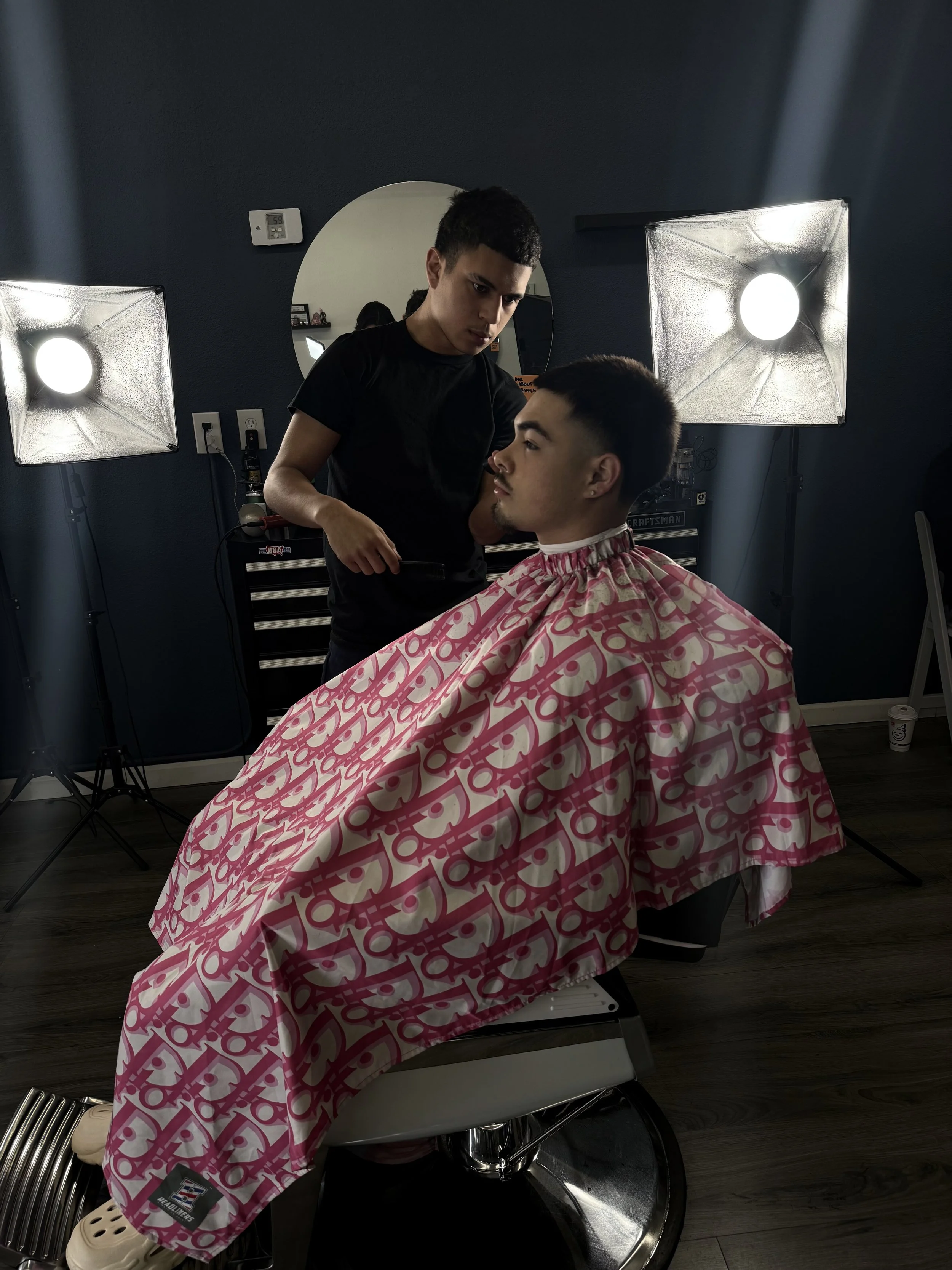 A young man gets a haircut at a barber shop, seated in a chair with a pink and white cape around his neck, while the barber prepares to cut his hair.