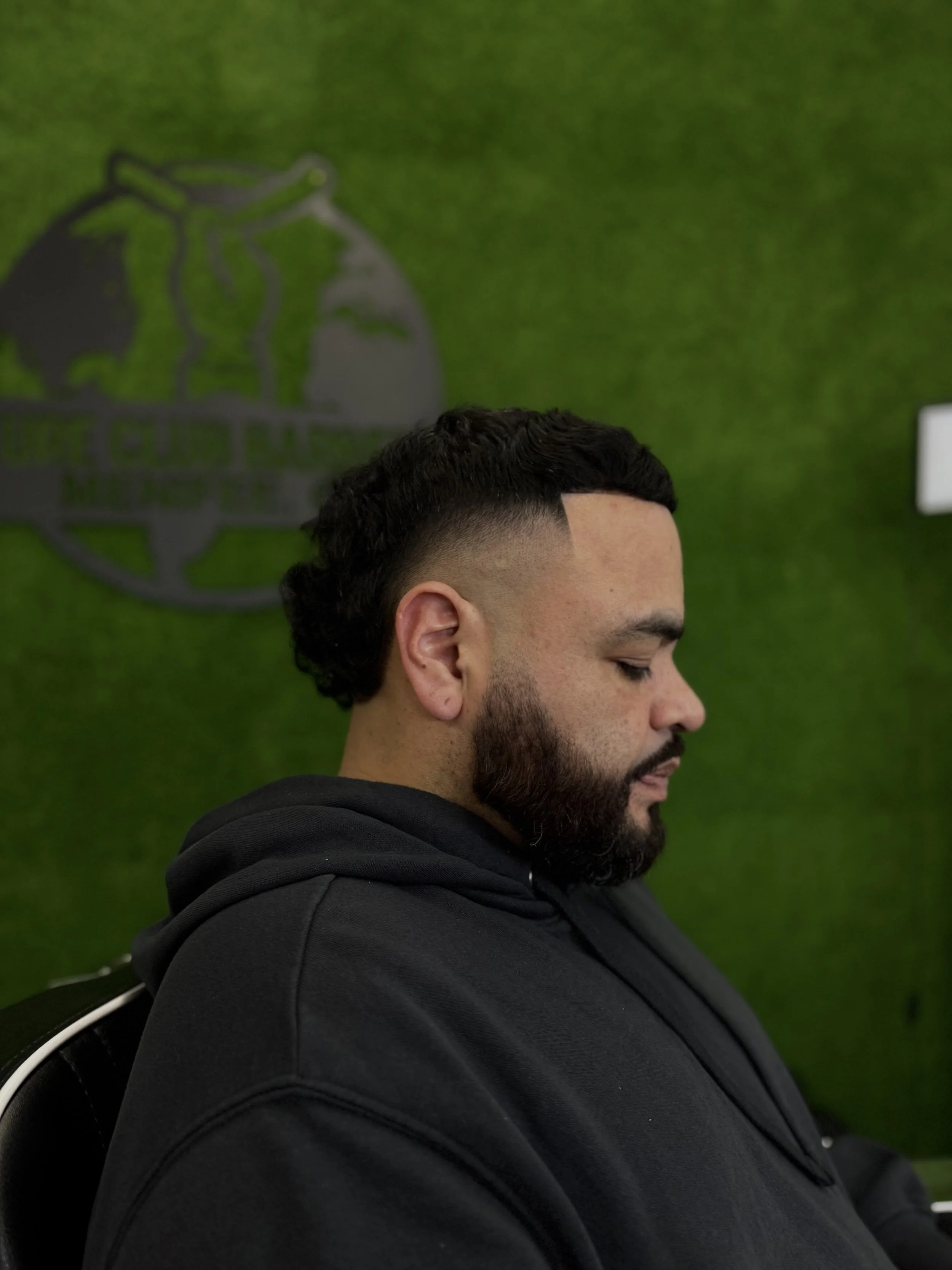 Side profile of a man with a fade haircut and a beard, sitting in front of a green wall with a logo. He is wearing a black hoodie and has his eyes closed.