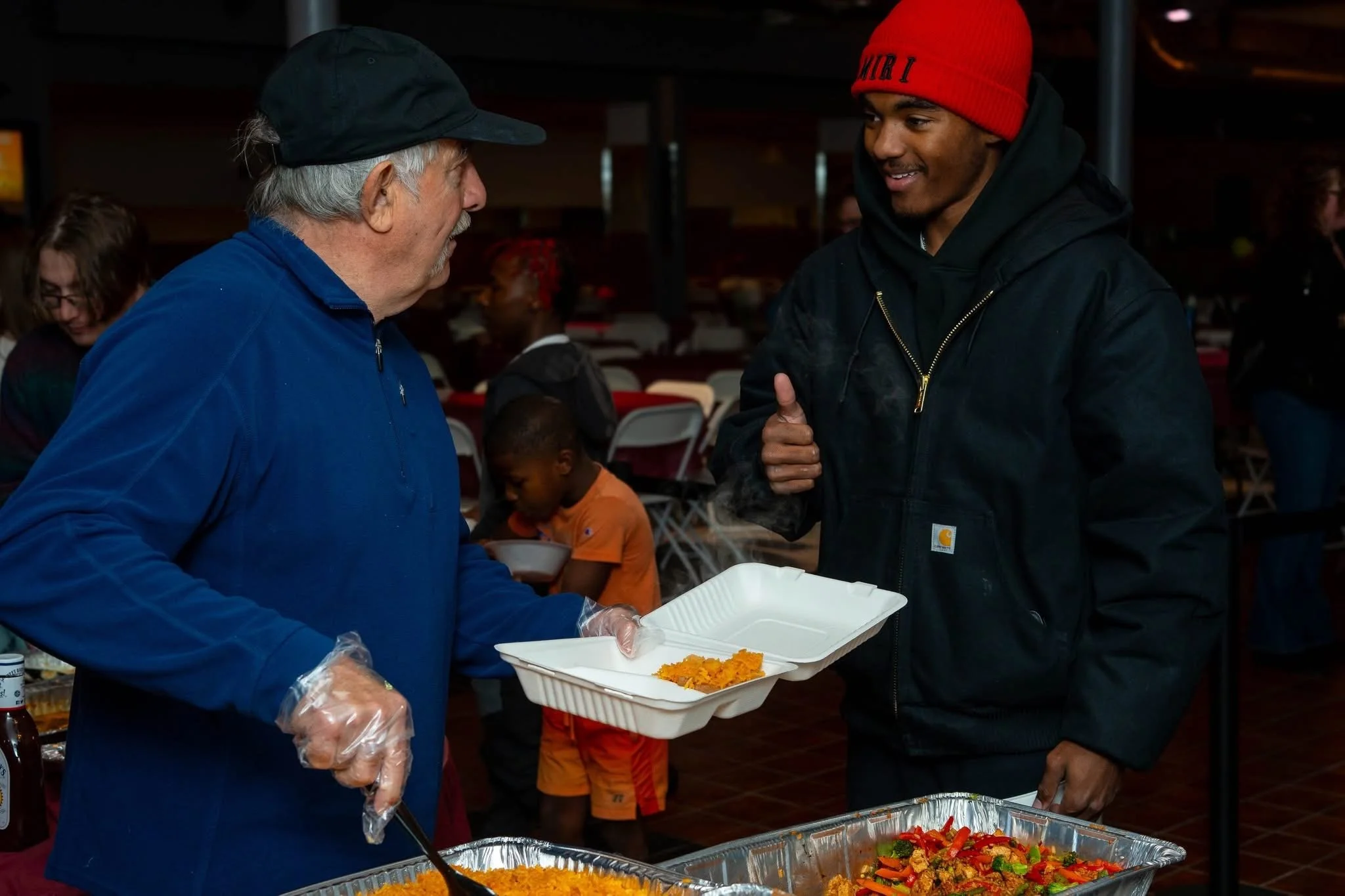 Two men, one older with gray hair wearing a blue jacket and hat, the other younger with a red beanie and black jacket, are shaking hands in a food serving area. A woman and child are in the background, and food trays with rice and vegetables are visible.
