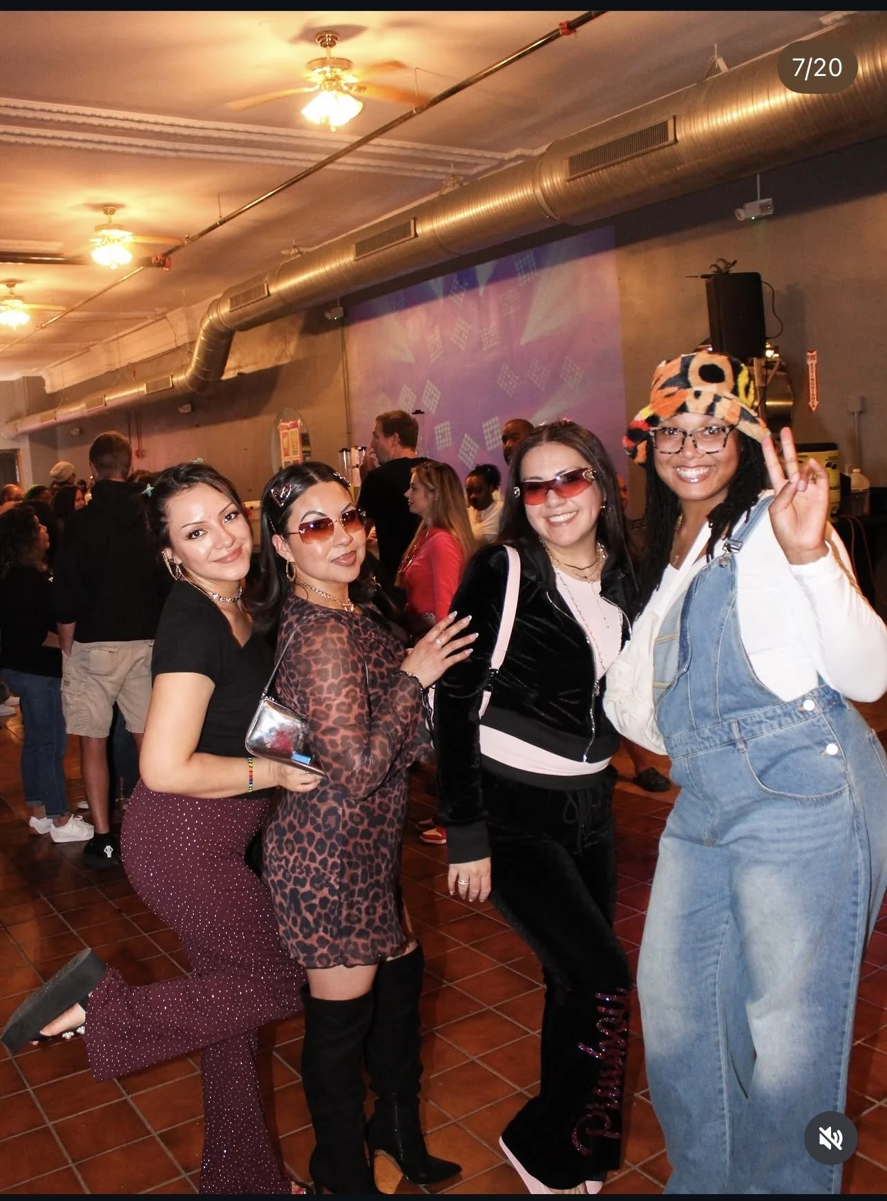 Four women posing for a photo at an indoor social event. They are dressed stylishly, with accessories and sunglasses, and are smiling. The background shows a festive atmosphere with other people mingling and a colorful light projection on the wall.