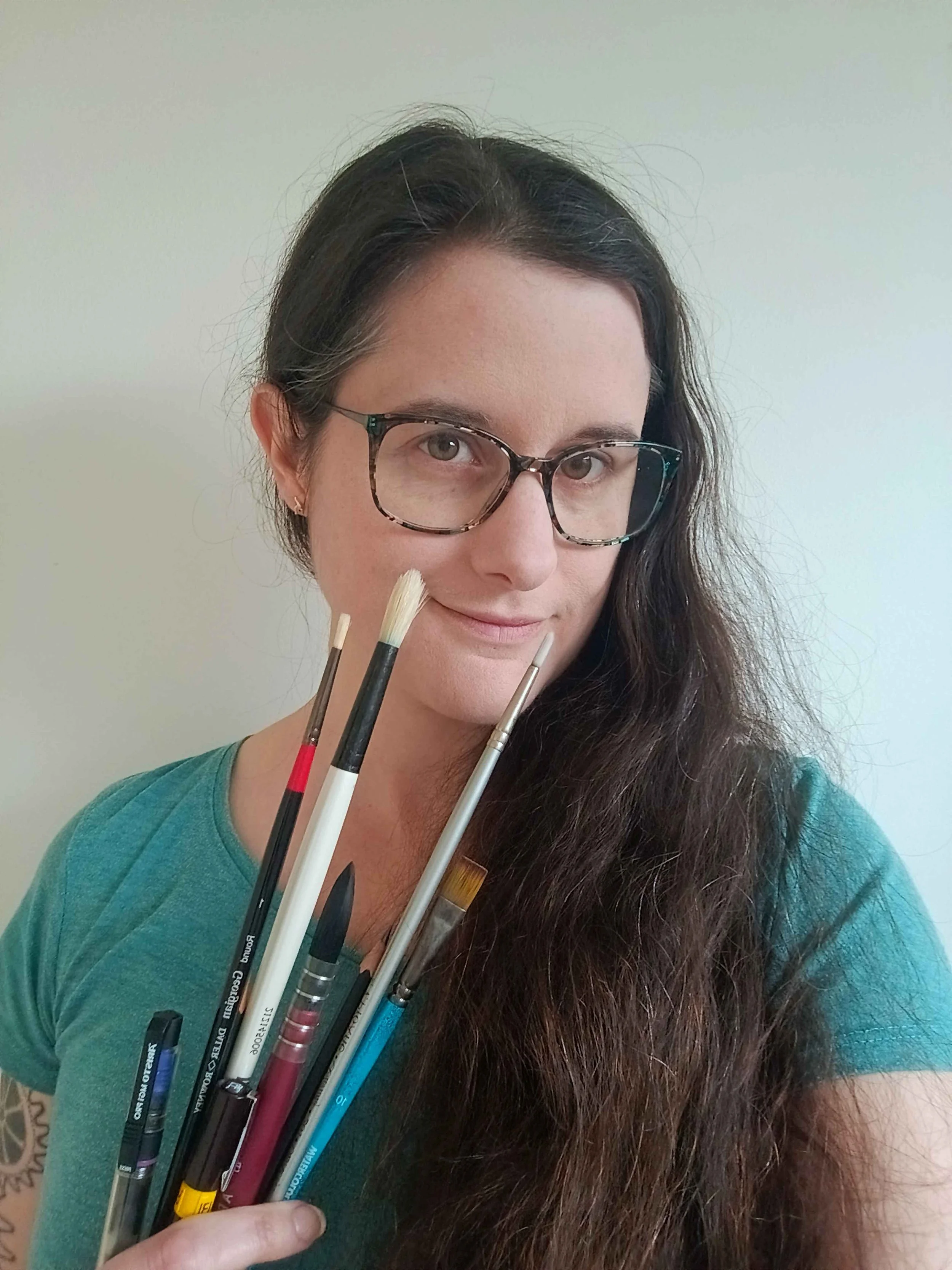 A woman with long dark hair, glasses, and a teal shirt, holding paintbrushes and art tools.