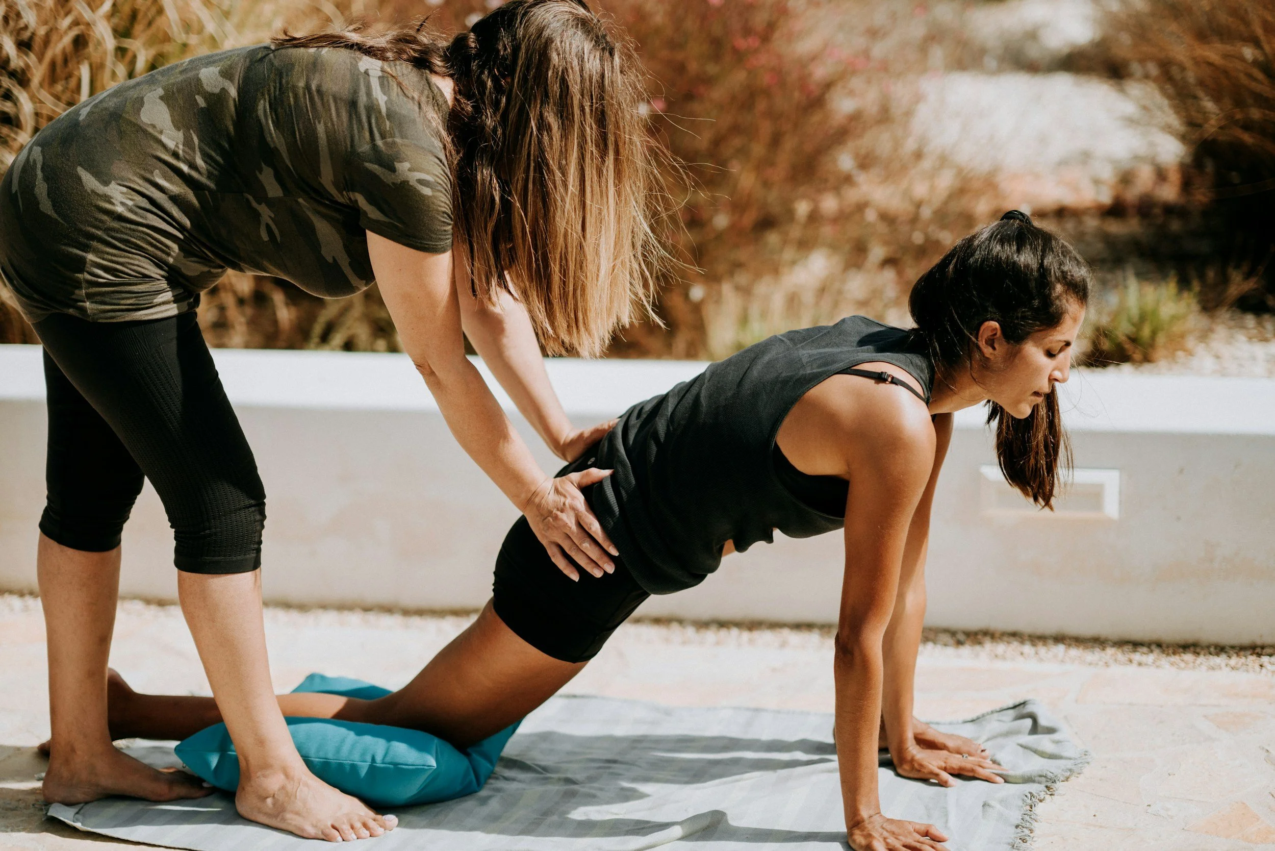 A woman doing a yoga plank position outdoors on a mat with assistance from another woman.