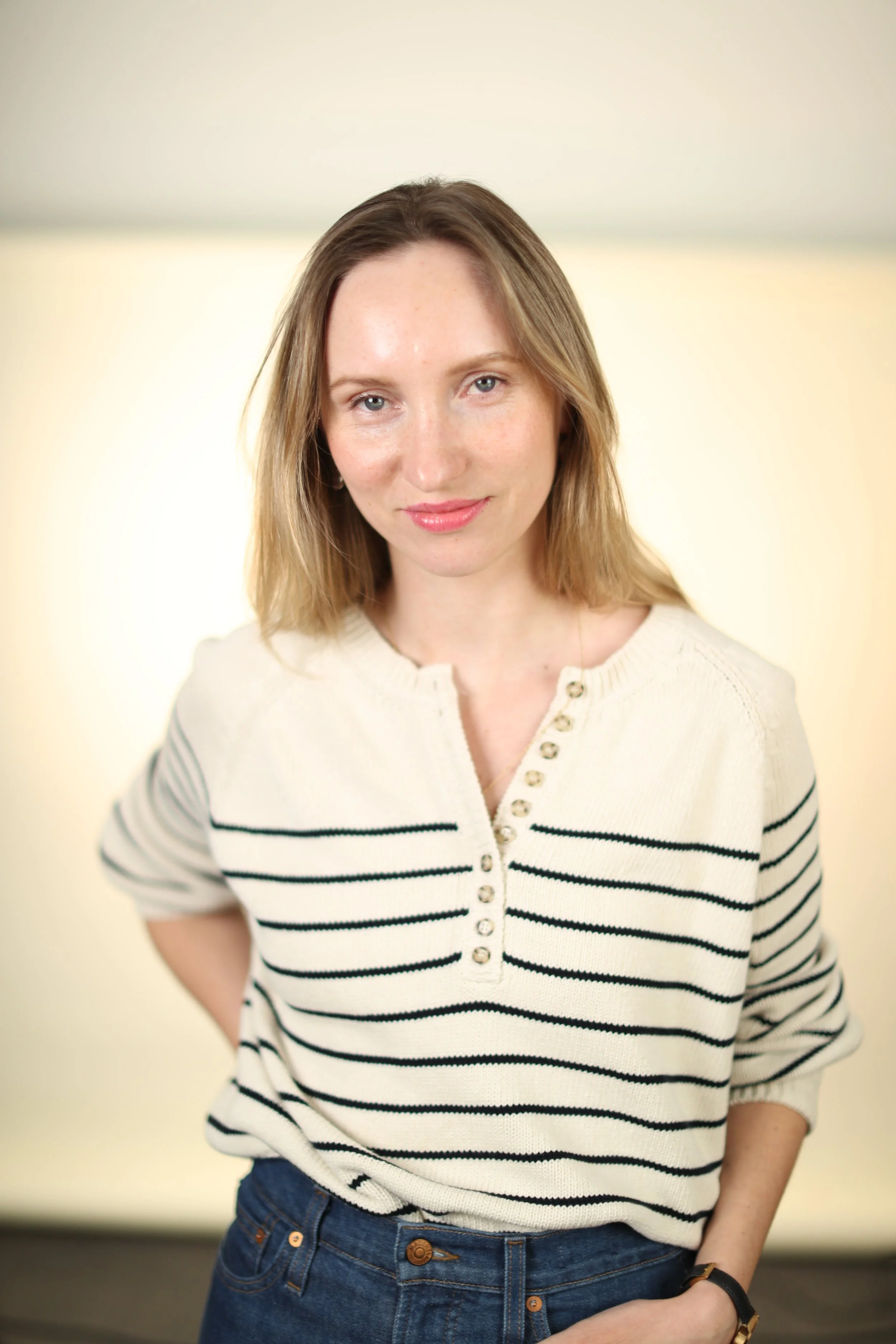 A woman with shoulder-length light brown hair, wearing a cream-colored sweater with black horizontal stripes and buttons, stands with her hands behind her back in front of a plain, light-colored background.