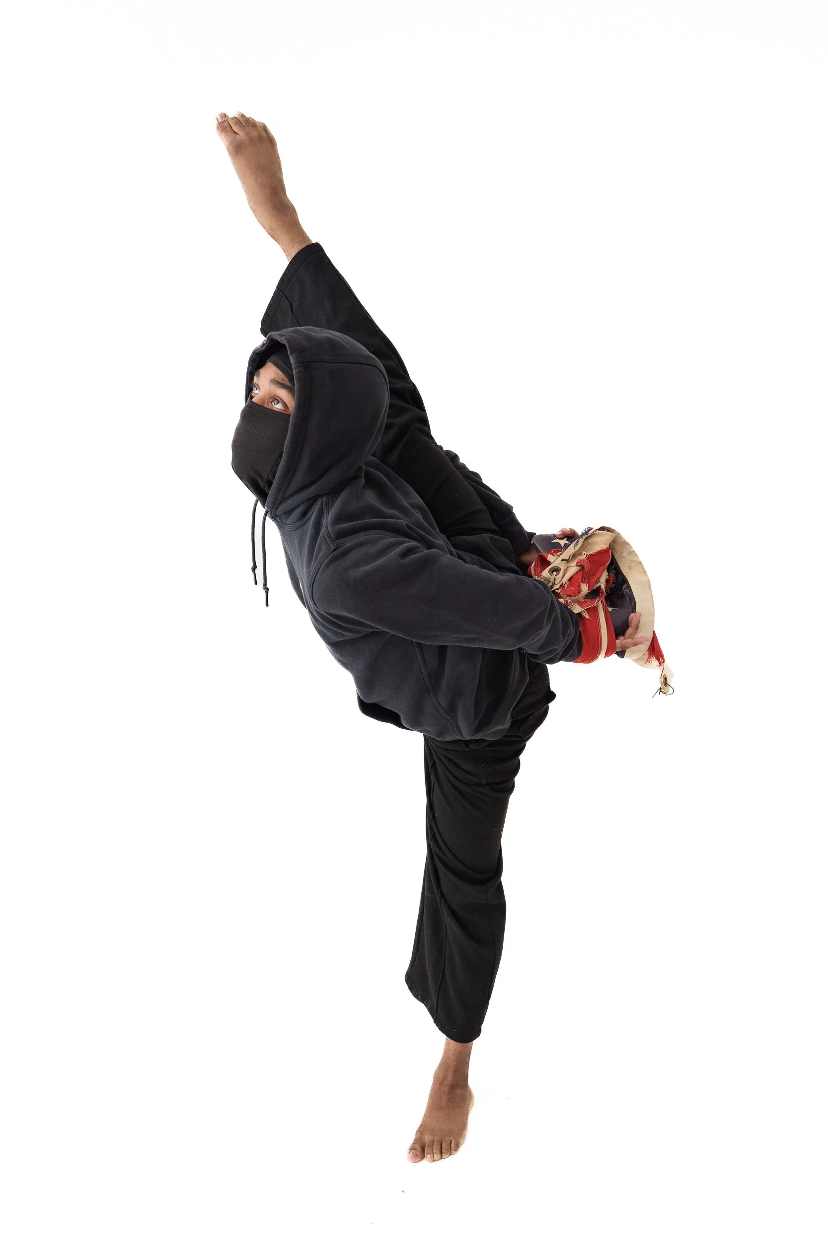 A young black male holds a pose: standing on left leg with right leg raised. His hands are tied behind his back with an American flag. He is looking upward and wearing all black for with a hood and face covering.