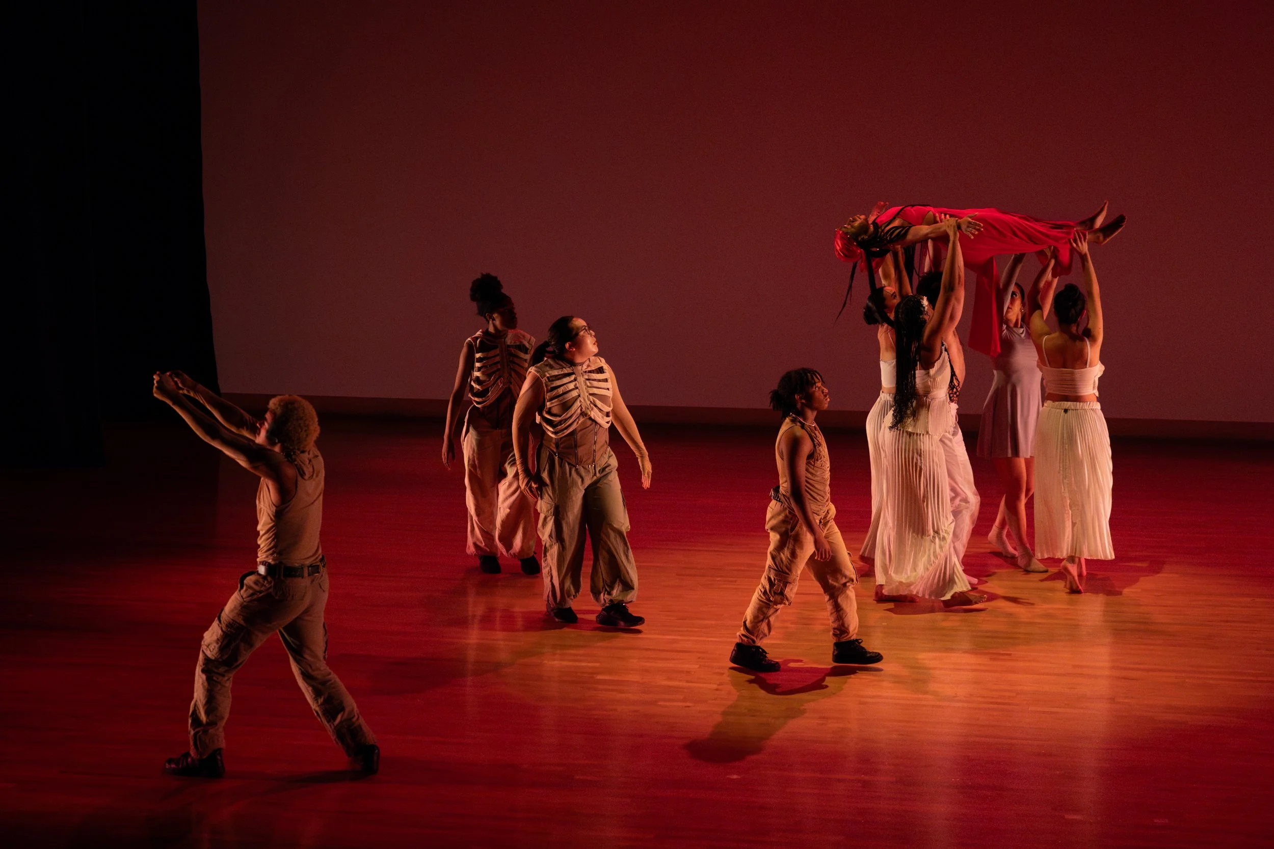 A group of dancers on stage, some lifting Aquilah Ohemeng professionally known as "KHILA" in a red dress, with a warm, red-toned backdrop.