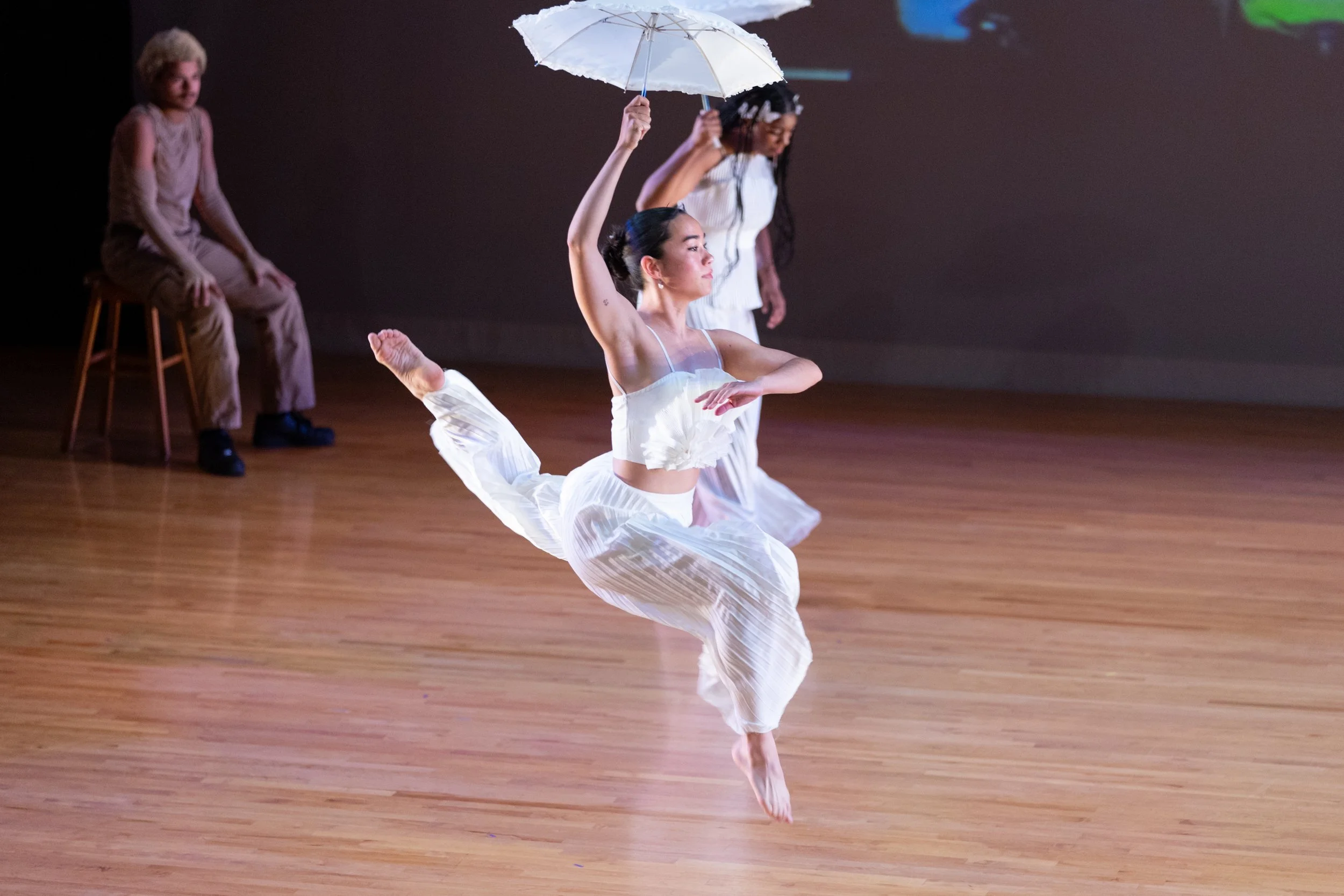 A woman leaps in the air holding a white umbrella on stage, dressed in white flowing clothing, while two other dancer sit in the background observing.