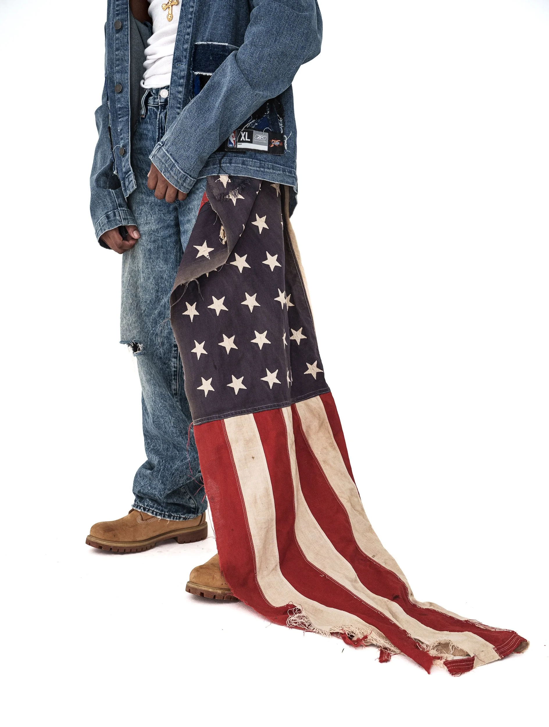 Part of a person wearing distressed denim jeans, a denim jacket, a white T-shirt with a gold cross necklace, and wide-leg pants with an American flag pattern, all standing against a white background.