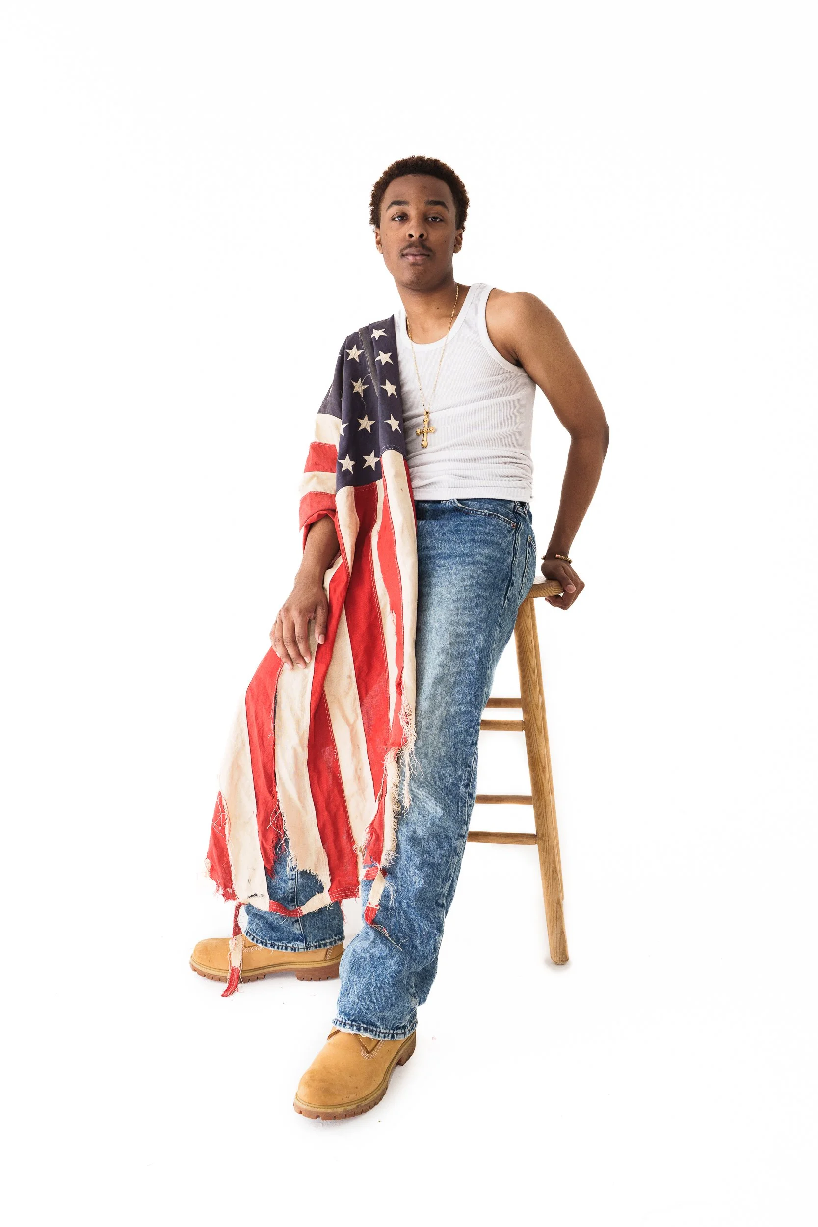 Young Black man with short curly hair wearing a white tank top, blue jeans, tan boots, and gold accessories, sitting on a stool with an American flag draped over his shoulder against a white background.