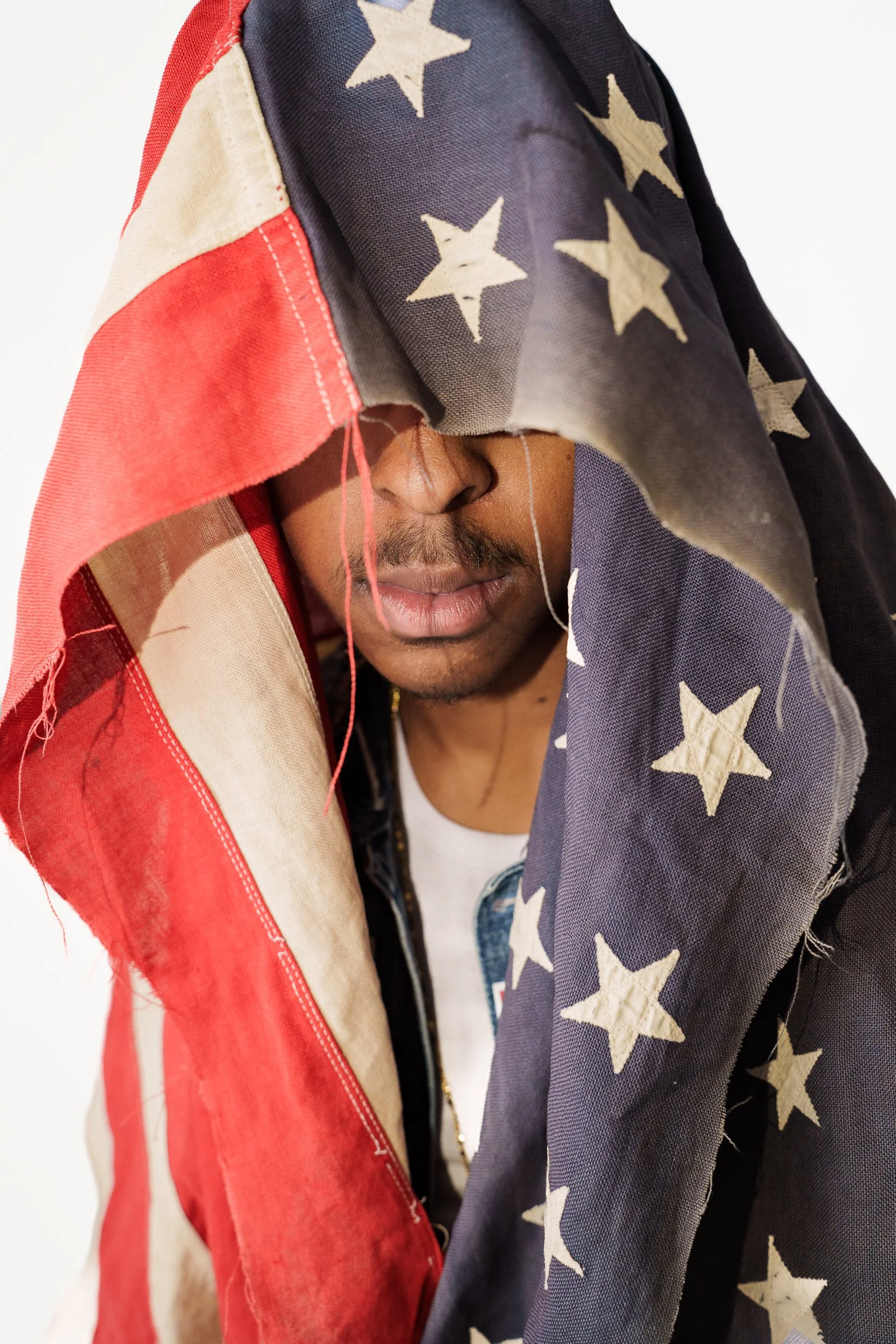 Close-up of a young, black male partially covered with a torn worn American flag with stars and stripes, draped over the head and shoulders for Aquilah Ohemeng professionally known as "KHILA"'s new project honoring Tupac Shakur