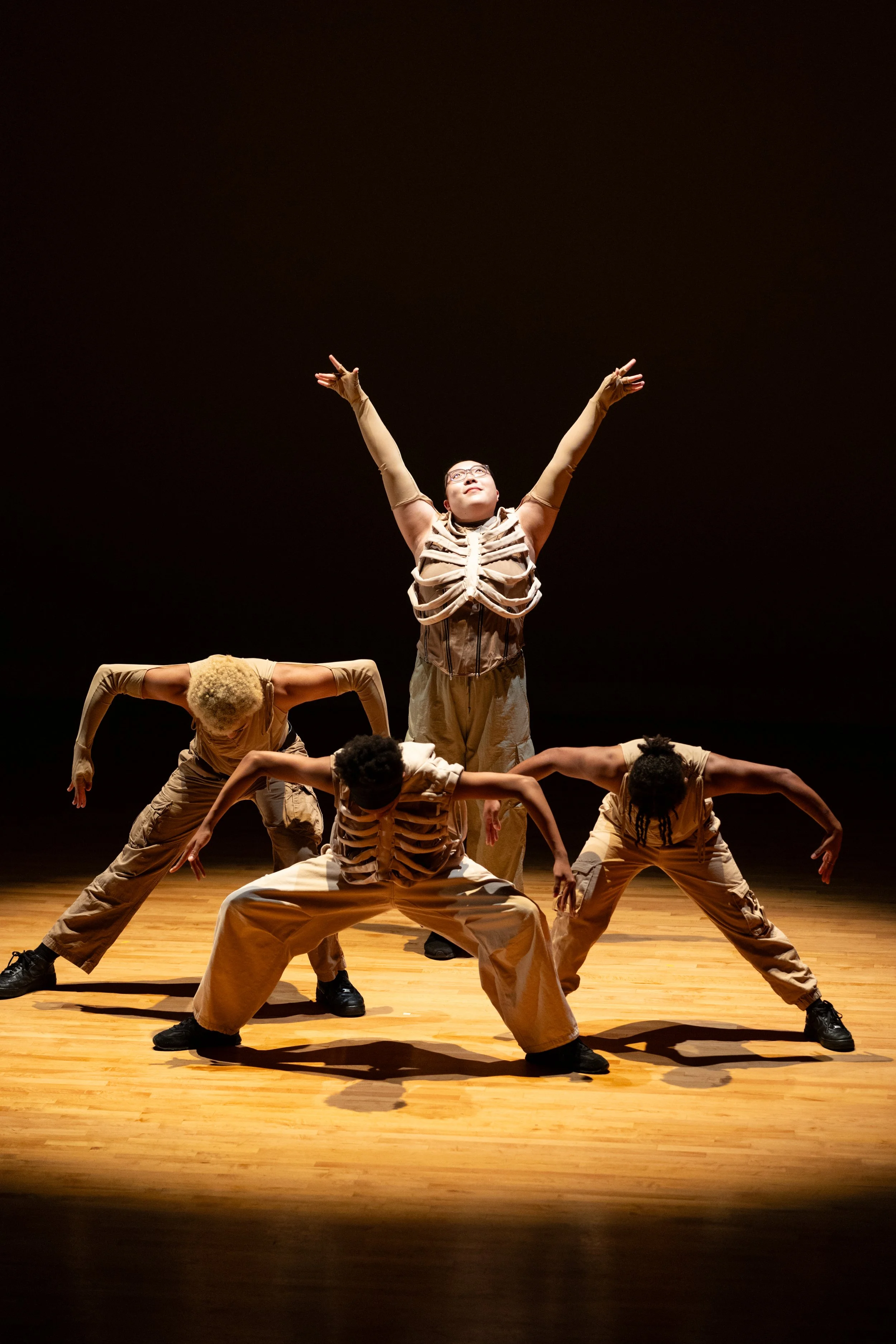 Group of dancers performing on stage, dressed in beige outfits with some wearing skeleton-themed costumes, striking various poses with arms and legs extended in a choreographed routine.