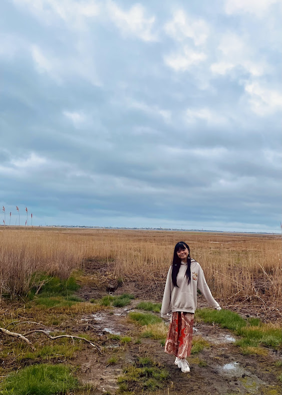 Young woman standing on a muddy trail in a grassy field with tall reeds under a cloudy sky during daytime.