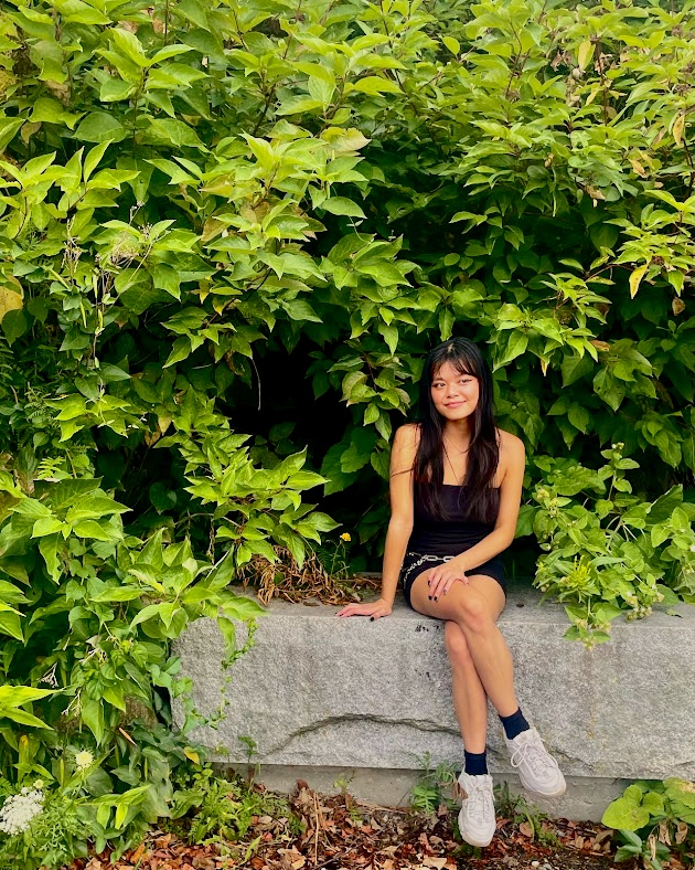 A young woman in a black dress sitting on a large stone surrounded by lush green foliage.