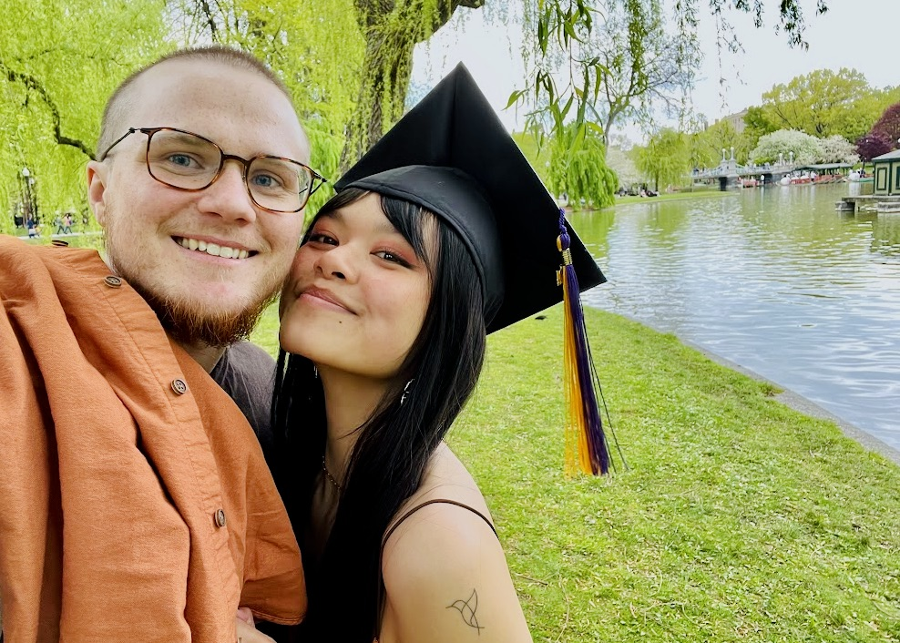 A smiling young woman in a graduation cap and gown taking a selfie with a smiling young man outdoors near a lake and trees.