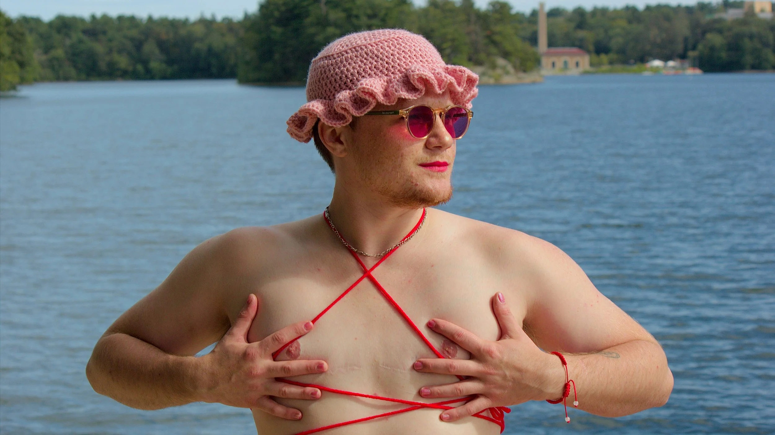 A white trans man wearing a pink crochet hat and tied with red yarn with hands on top surgery scars against a lake backdrop in Massachusetts