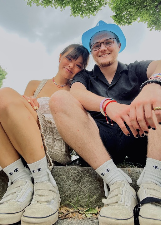 Two young people sitting outdoors under a tree, smiling. The girl is wearing beige shorts, white socks, white sneakers, and a necklace. The boy is wearing black shorts, a black shirt, glasses, and a blue hat, with several bracelets on his wrist.