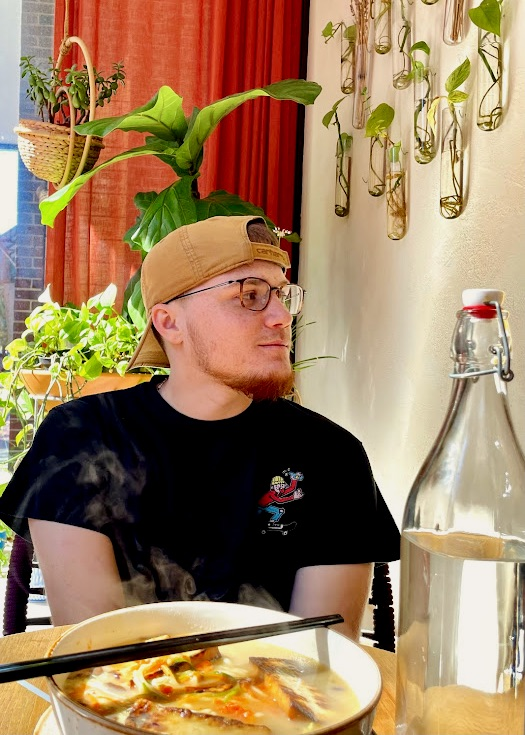 Young man with glasses and a beige cap sitting at a table with hot ramen, surrounded by green plants and a white wall with hanging glass planters.
