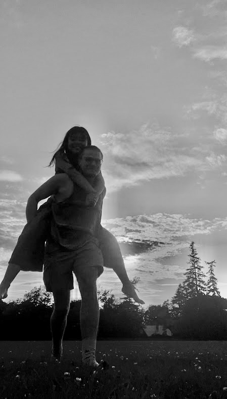 A person giving a piggyback ride to a child outdoors during sunset, with trees and a cloudy sky in the background.
