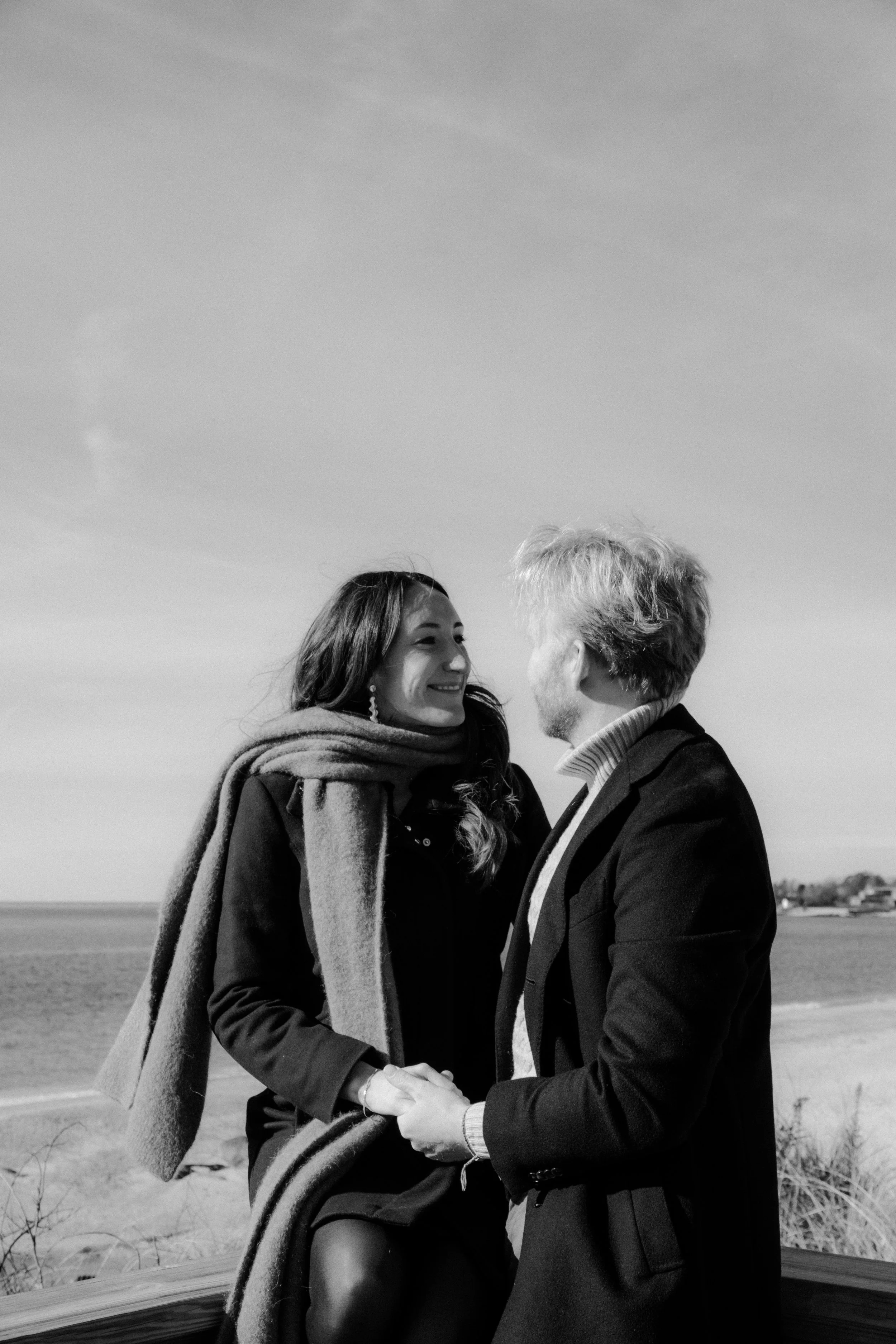 A black and white photo of a couple holding hands and smiling at each other on a beach, with the ocean in the background and a clear sky.