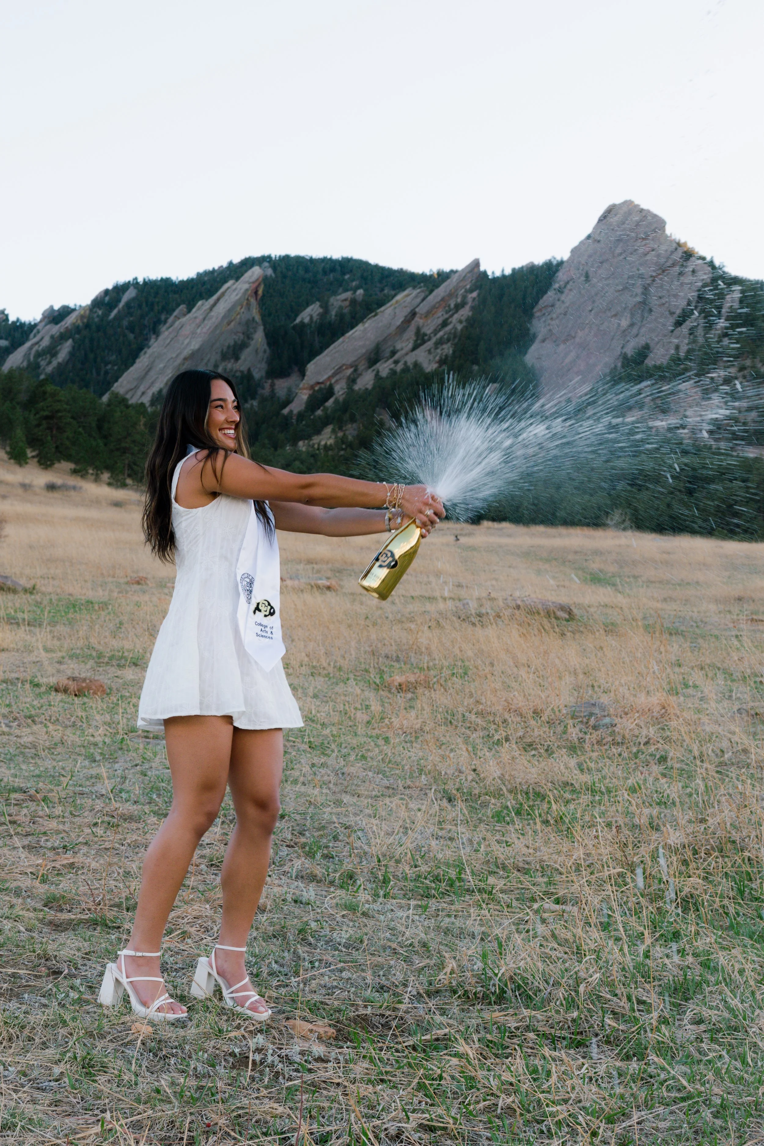 Woman in white dress and high heels celebrating graduation by spraying champagne in an open field with mountains in the background.