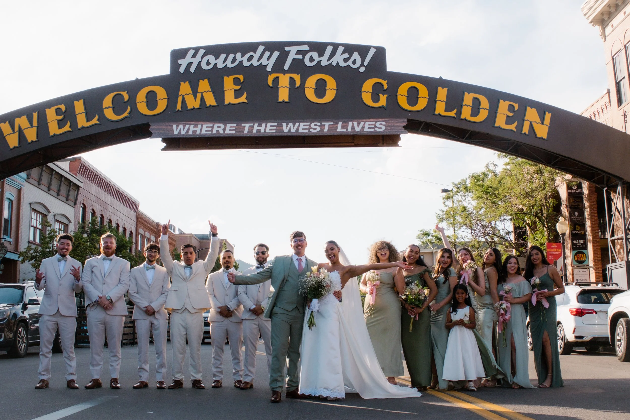 A wedding party standing under a welcoming arch that says "Howdy Folks! Welcome to Golden, where the west lives" on a city street with buildings and parked cars.