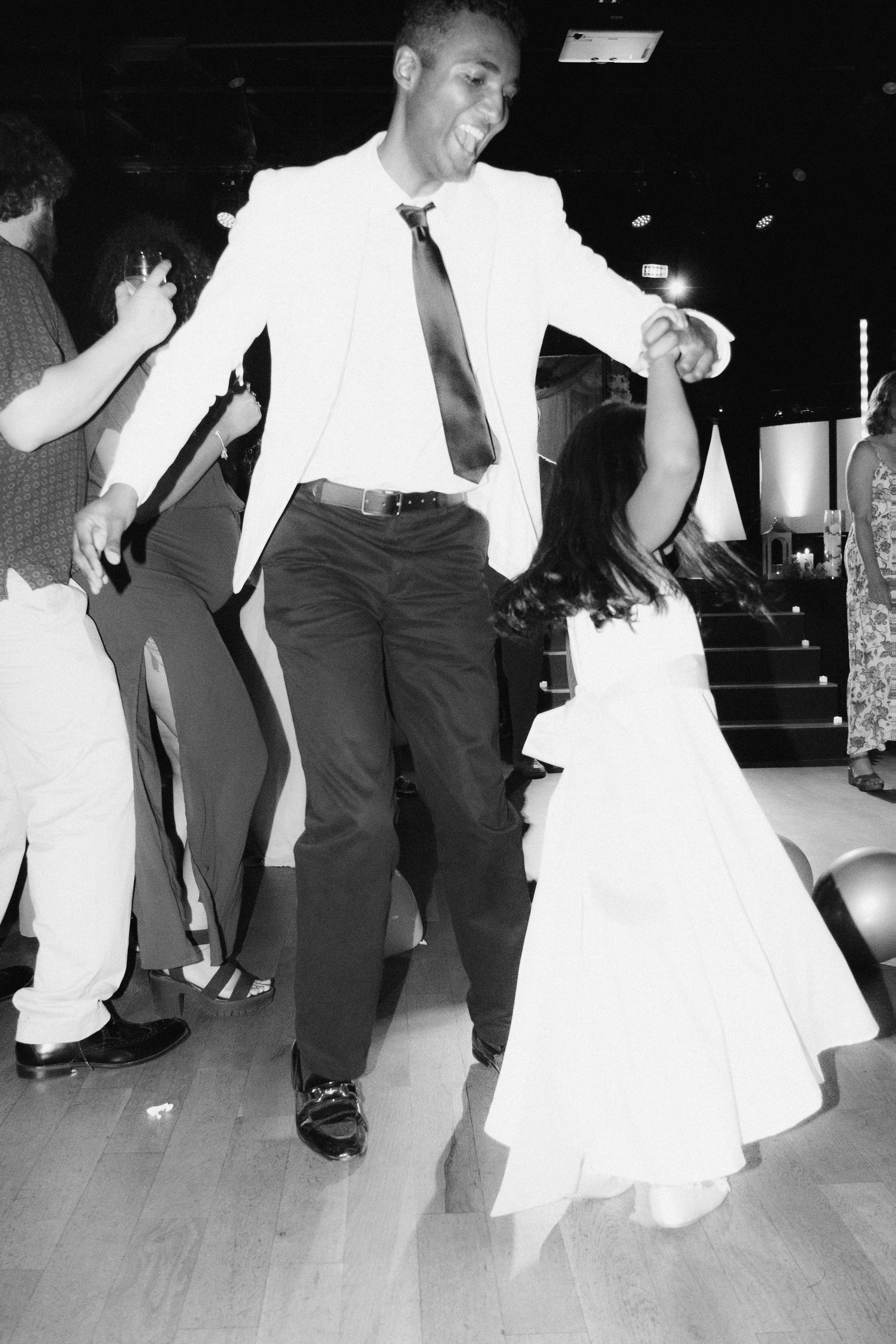 A man in a white suit dancing with a young girl in a white dress at a celebration or party.