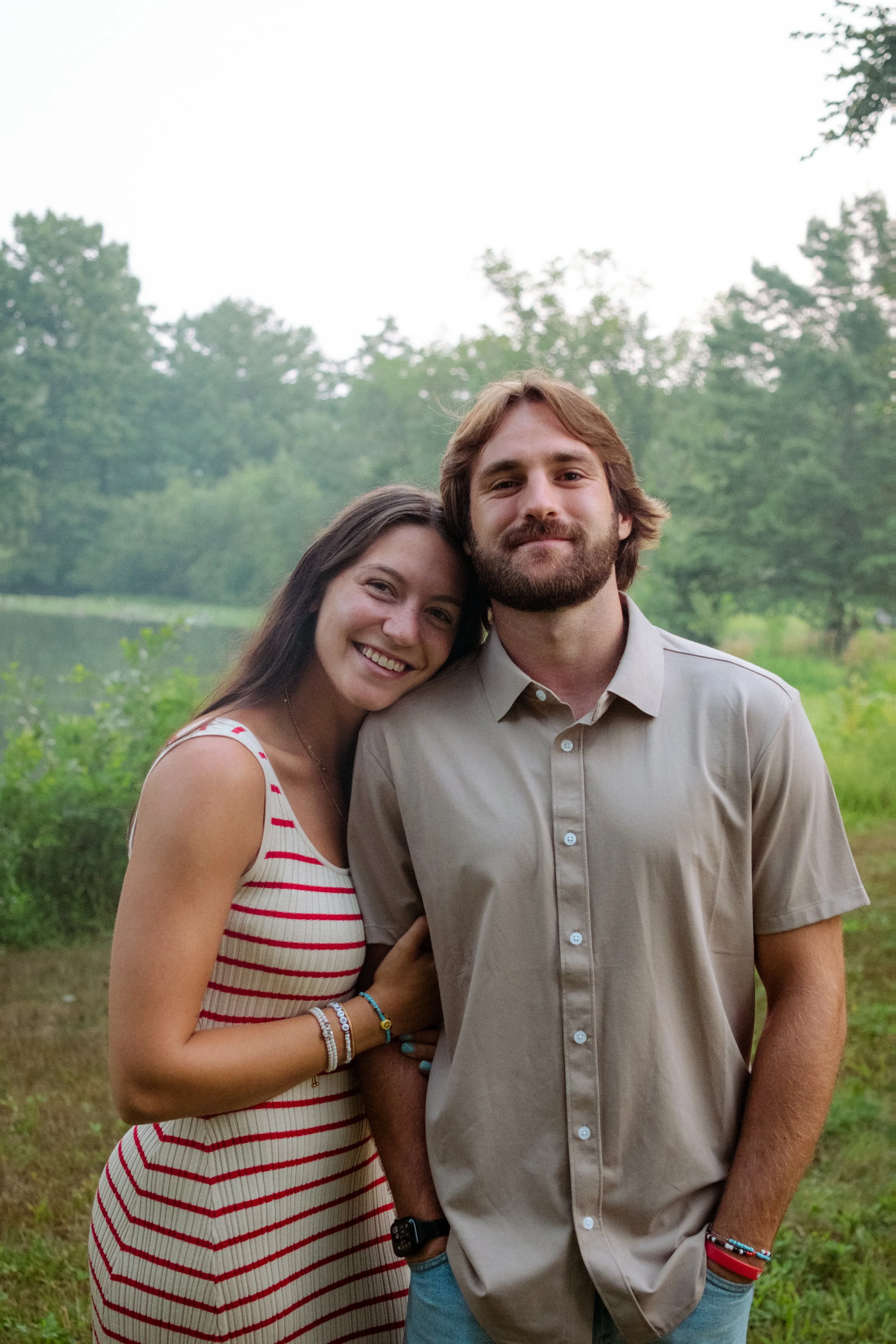 A young woman and man standing outdoors near a lake with trees in the background, smiling and closely posing together.
