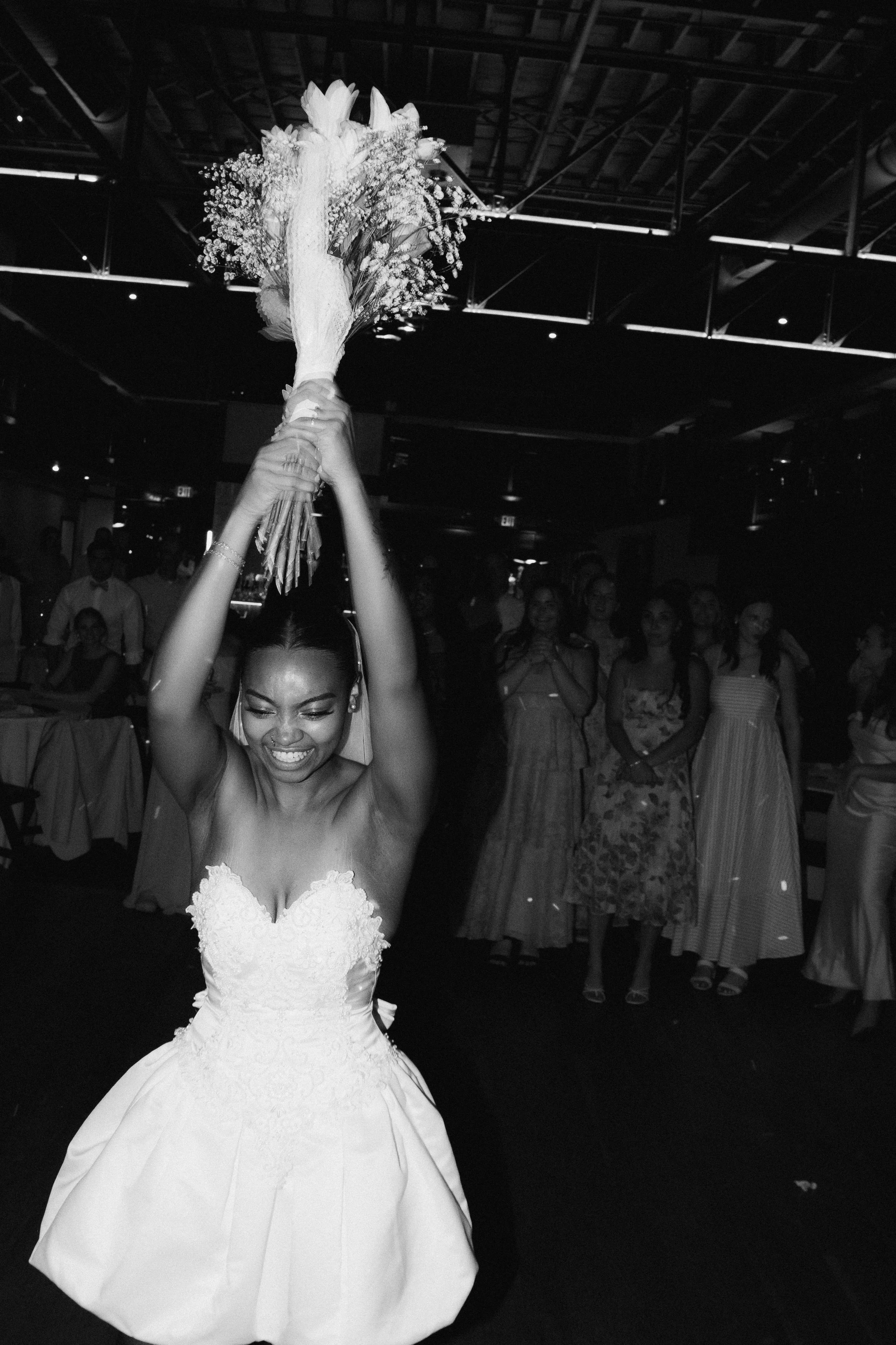 A woman in a wedding dress holding a bouquet of flowers above her head, smiling, at a wedding reception with guests in the background.