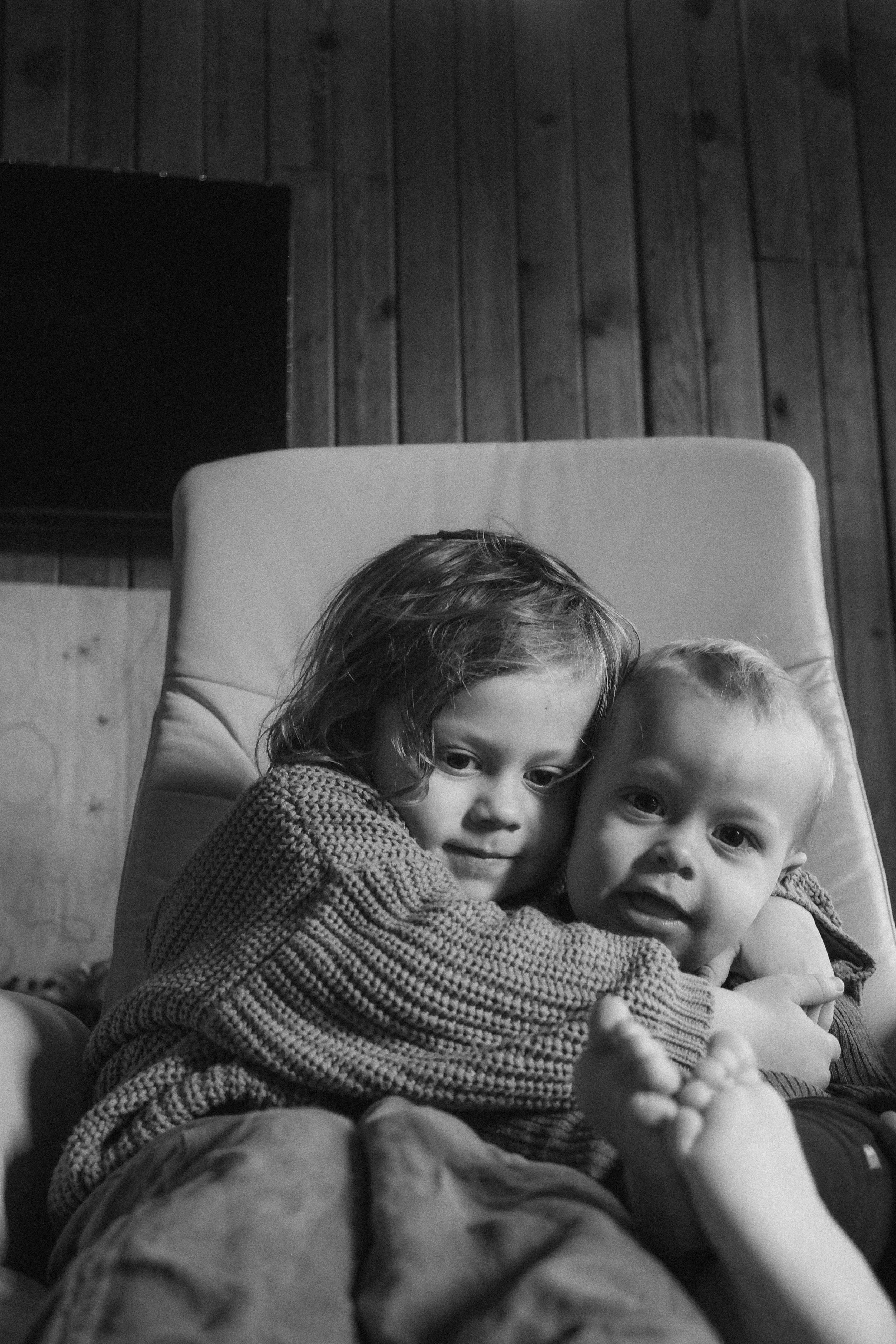 Two young children, a girl and a boy, sitting on a couch hugging each other in a cozy room with wooden wall paneling, captured in black and white.