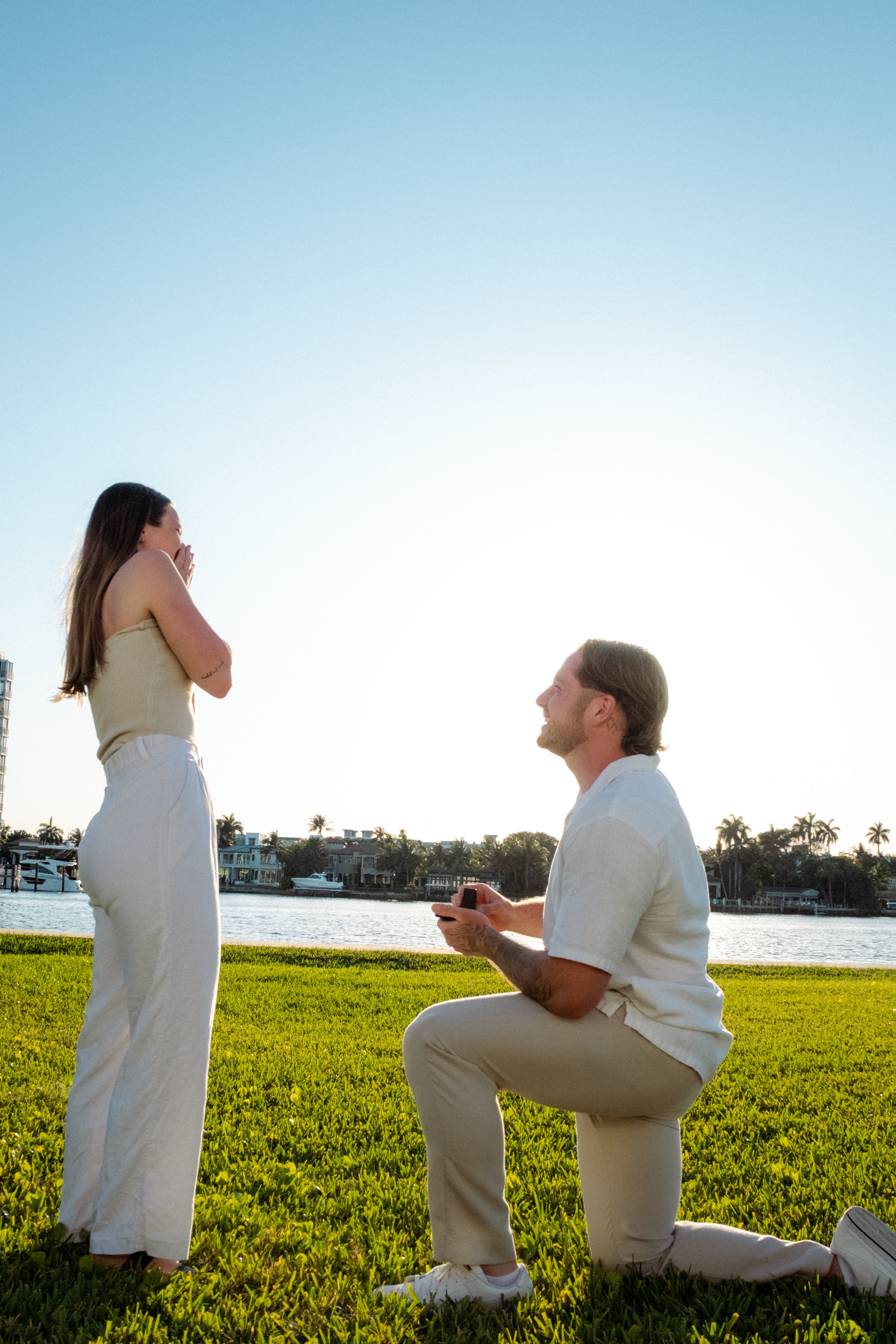 A man kneeling on the grass proposing to a woman by a body of water during sunset, with her appearing surprised and emotional.