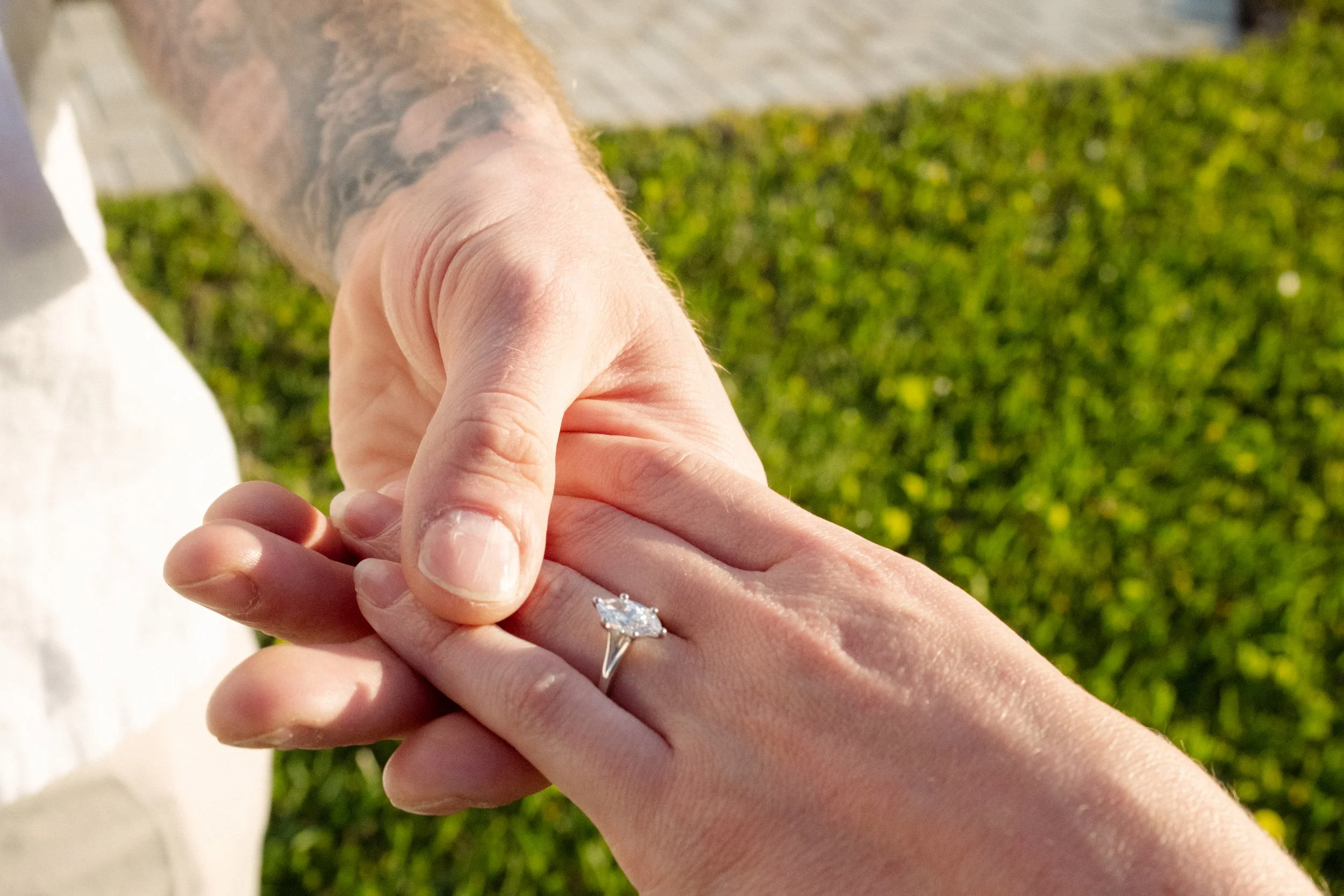 Close-up of a person's hand holding another person's hand, showing an engagement ring with a large diamond on the ring finger, with grass and sidewalk in the background.