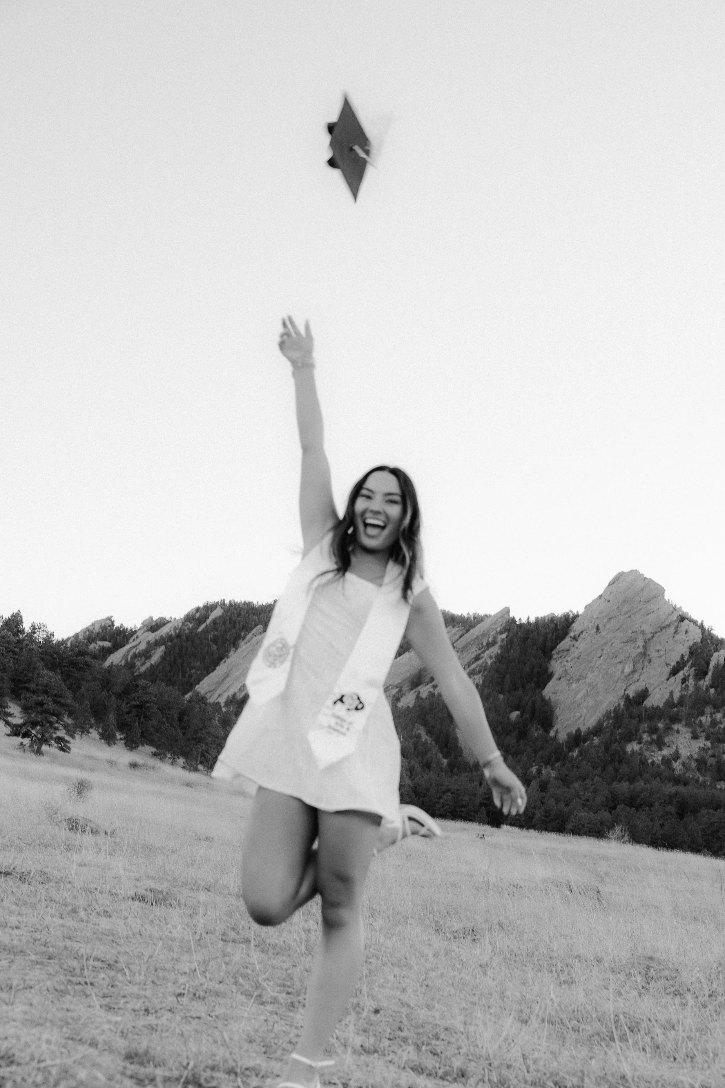 A black and white photo of a woman in a graduation gown celebrating outdoors in a field with mountains in the background. She is smiling and throwing her graduation cap into the air.