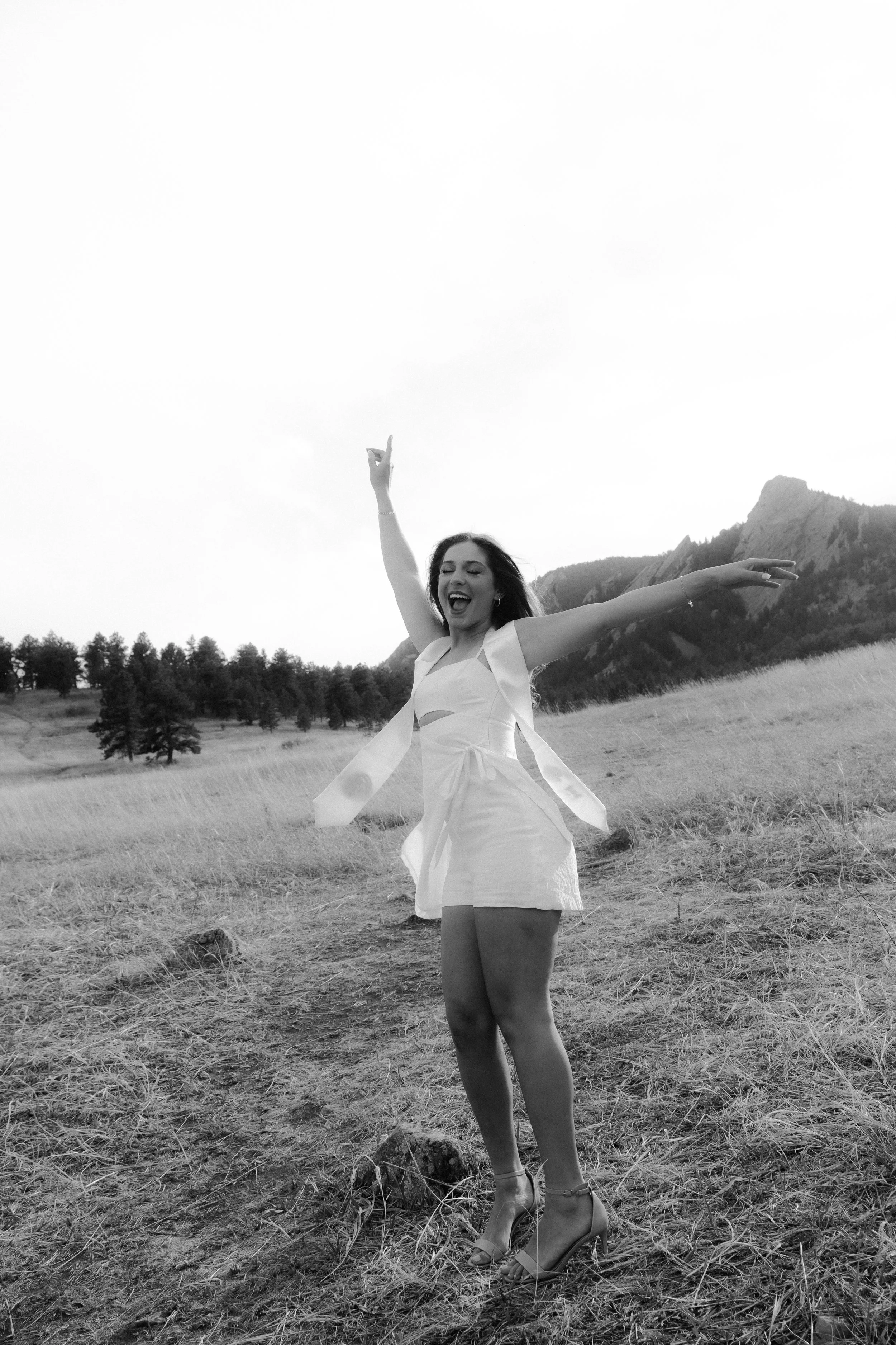 A woman in a white dress and high heels joyfully dancing in a field with mountains in the background, captured in black and white.