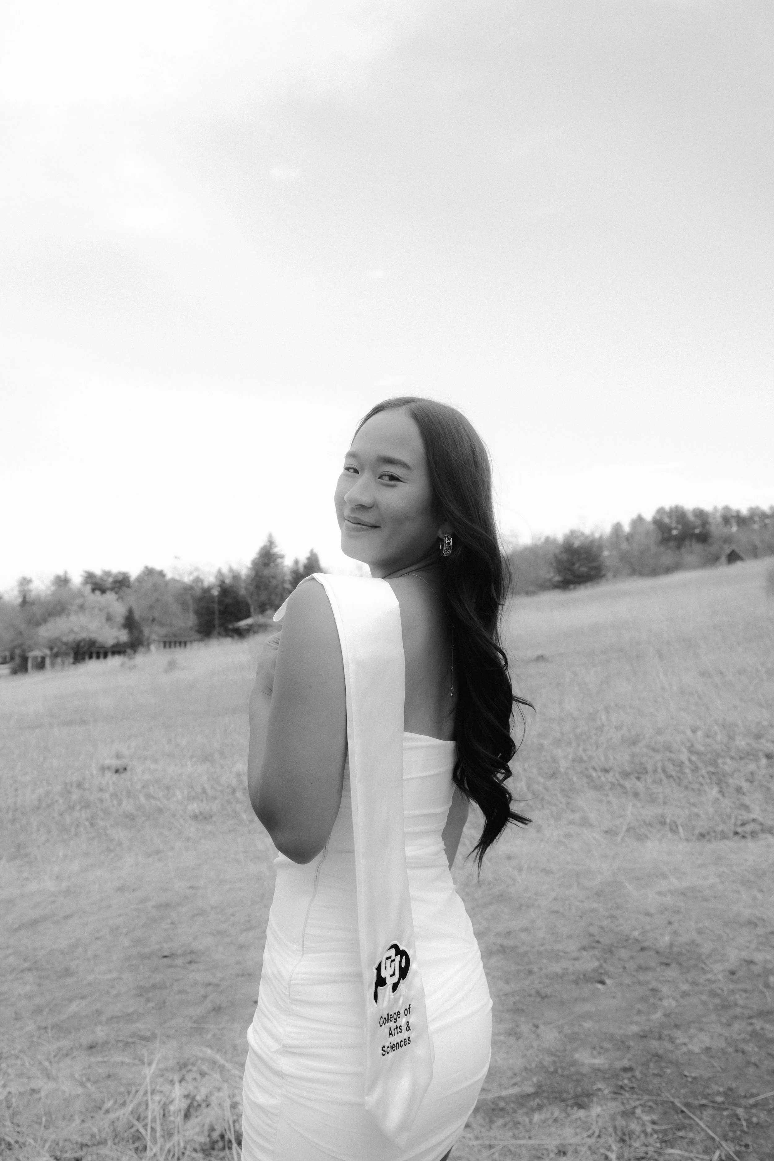 A smiling young woman in a white dress stands outdoors in a field, with trees and a house visible in the background on a clear day.
