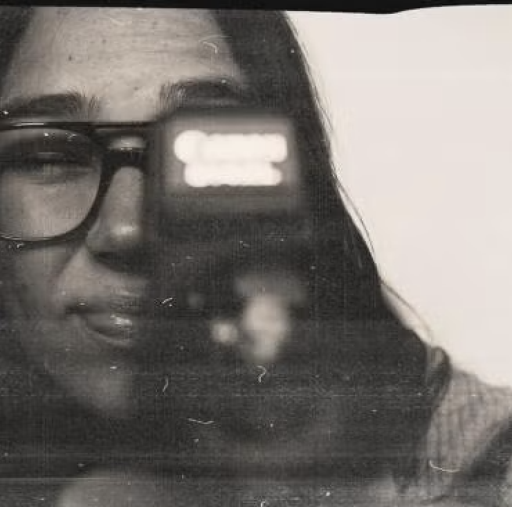 Black and white photo of a woman wearing glasses, with her face partially visible in a mirror reflection, holding a phone to take a selfie.