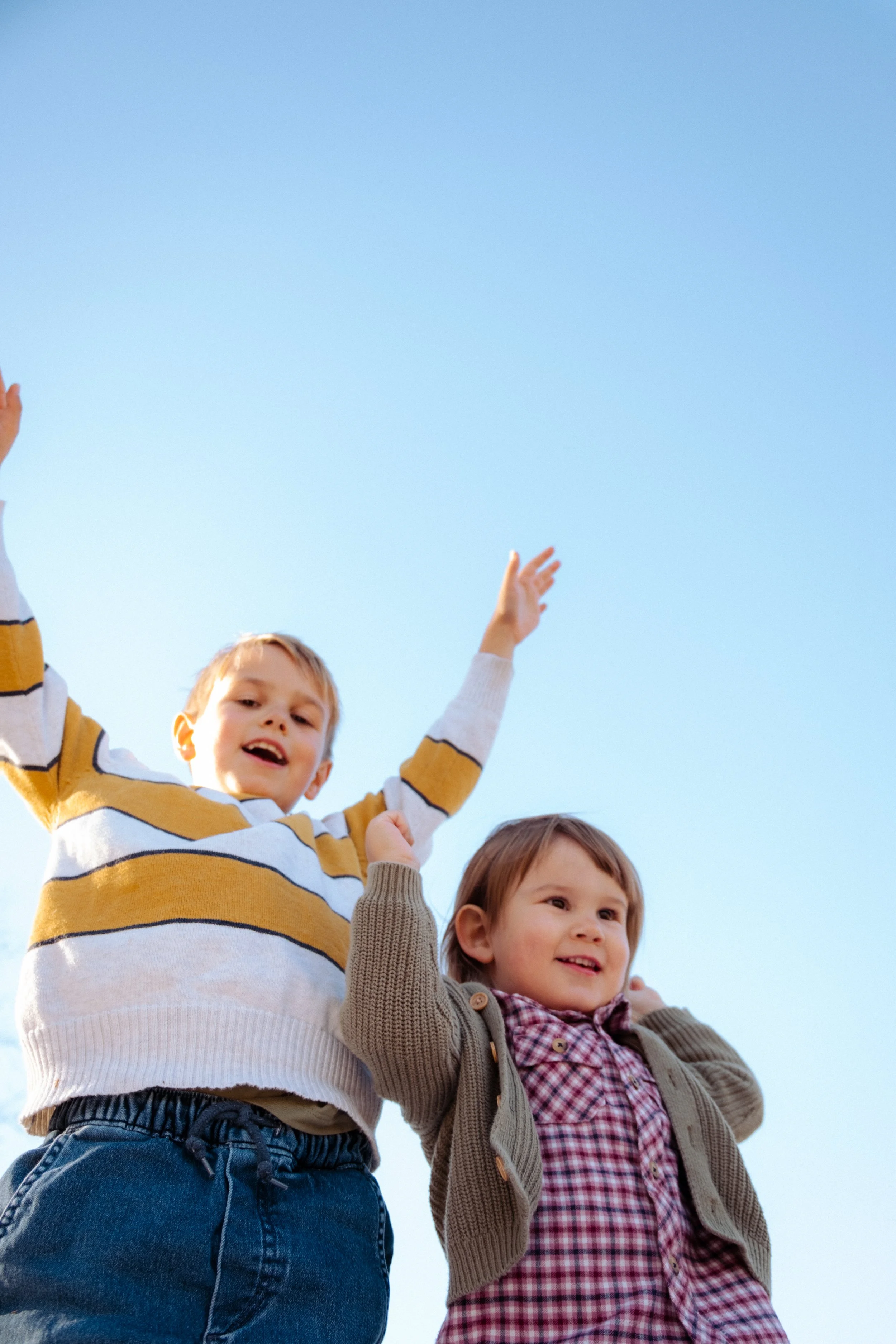 Two young children, a boy and a girl, looking up and smiling with their arms raised, against a clear blue sky.