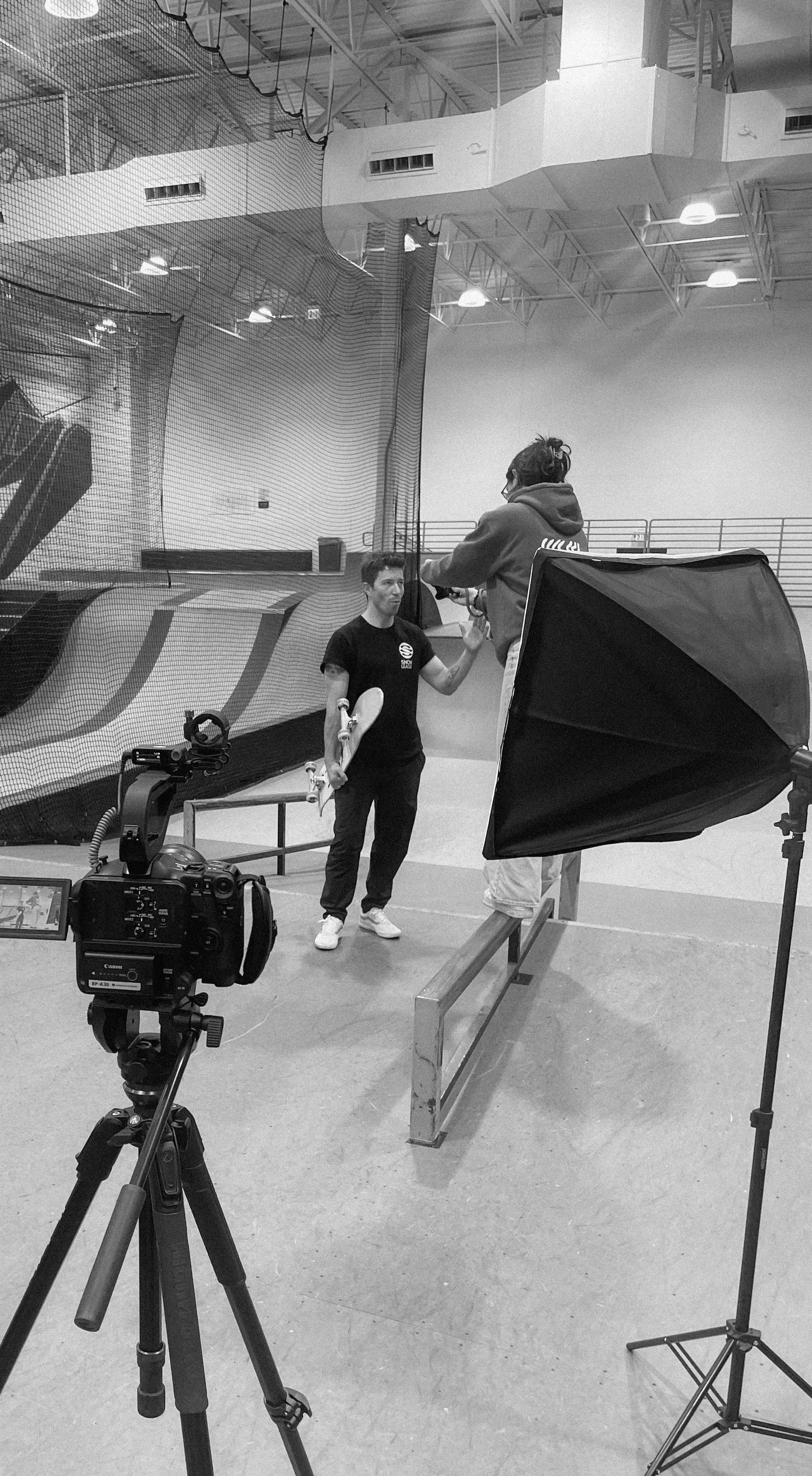 A skateboarding indoor facility with two individuals, one holding a skateboard, involved in a photo shoot. There is professional photography equipment including a camera on a tripod, a softbox, and a backdrop.