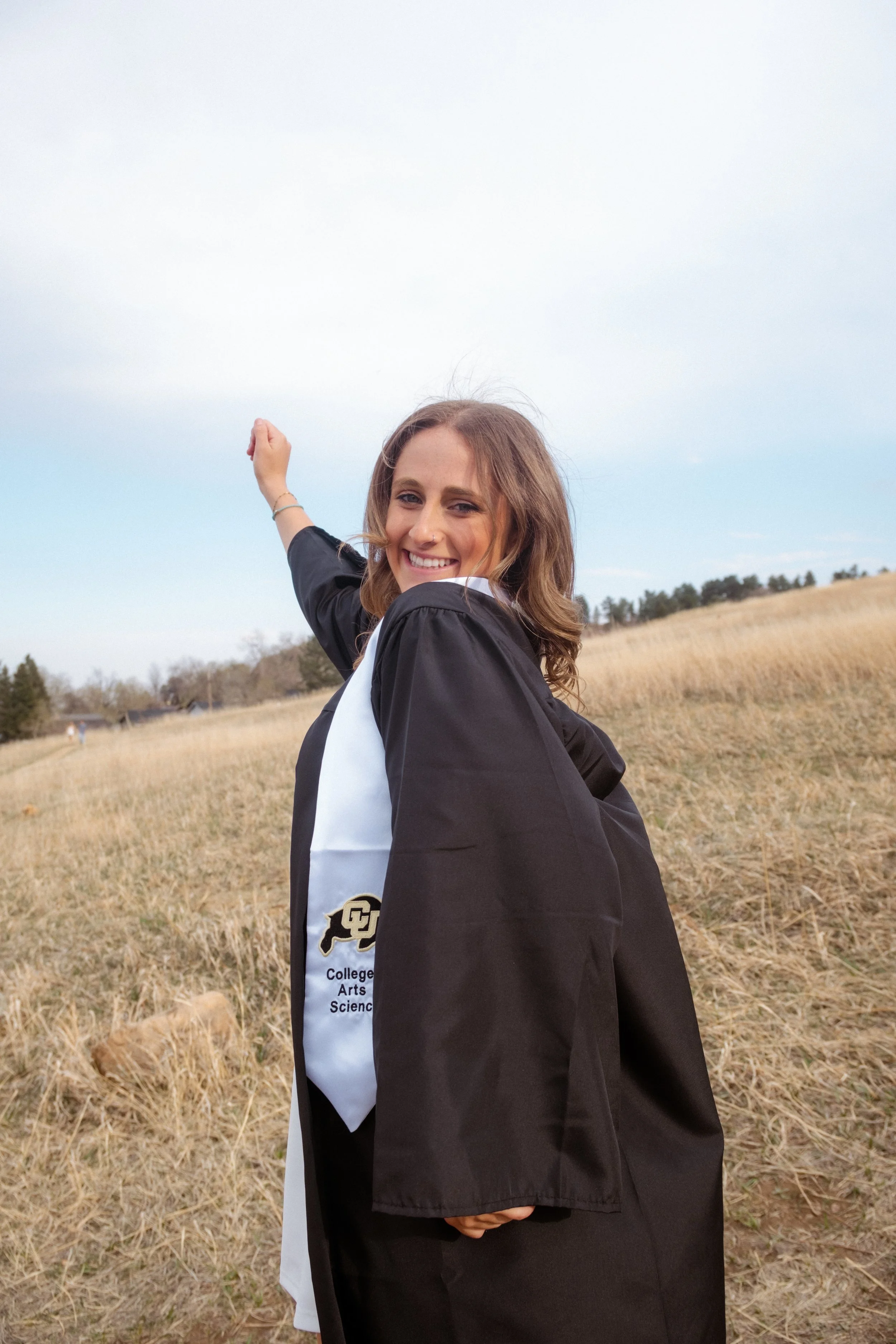 A young woman in a graduation gown and hood, smiling and pointing into the distance outdoors in a grassy field with a cloudy sky.