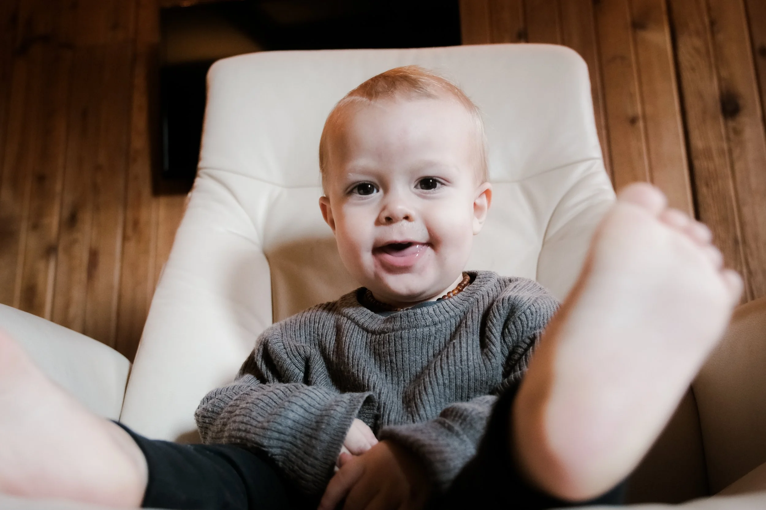A young child with light skin, red hair, and a brown necklace sitting in a white chair, smiling at the camera with one foot towards the lens, in a room with wooden walls.