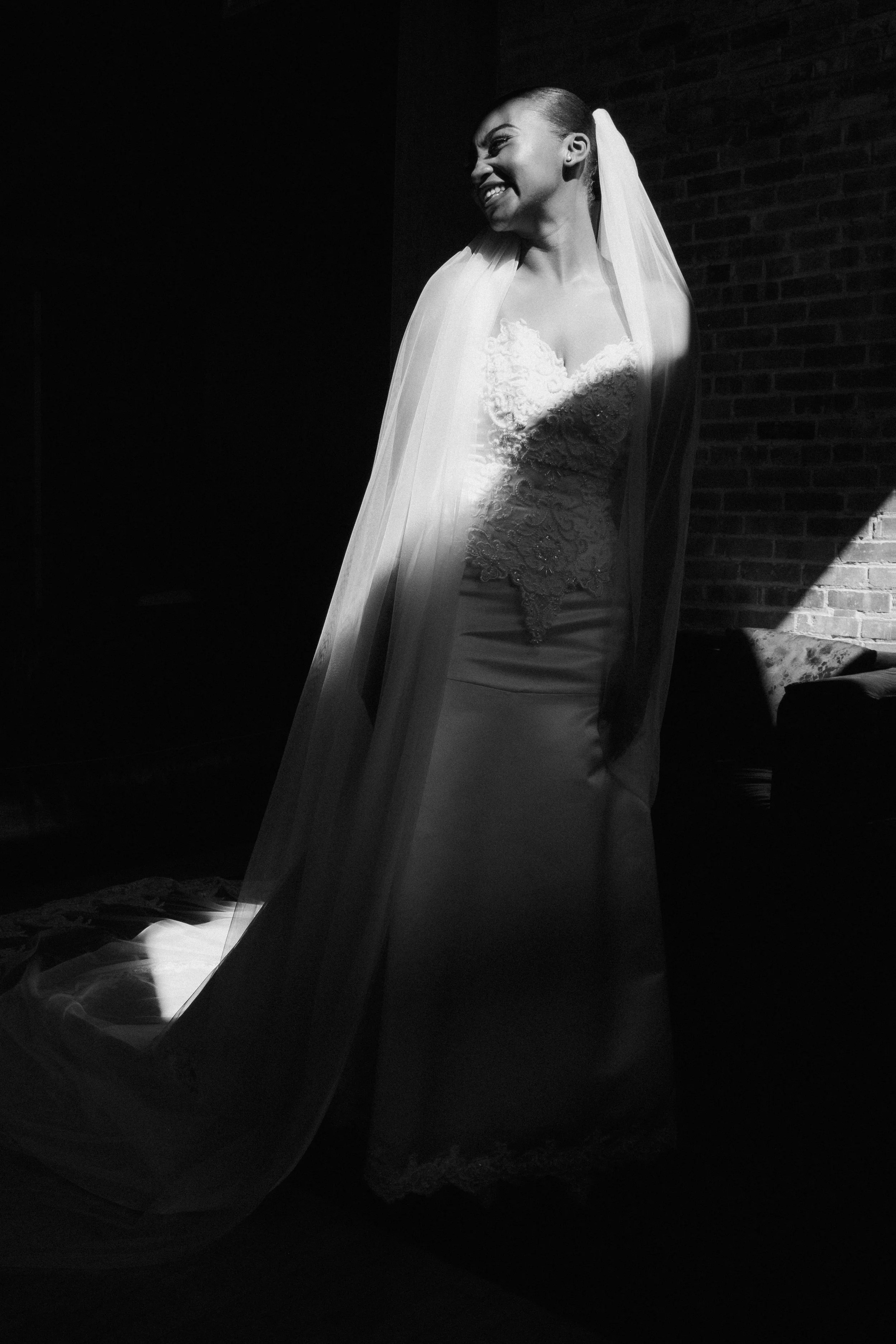 Black and white photo of a bride smiling, wearing a wedding dress and veil, standing against a brick wall with a dramatic shadow.