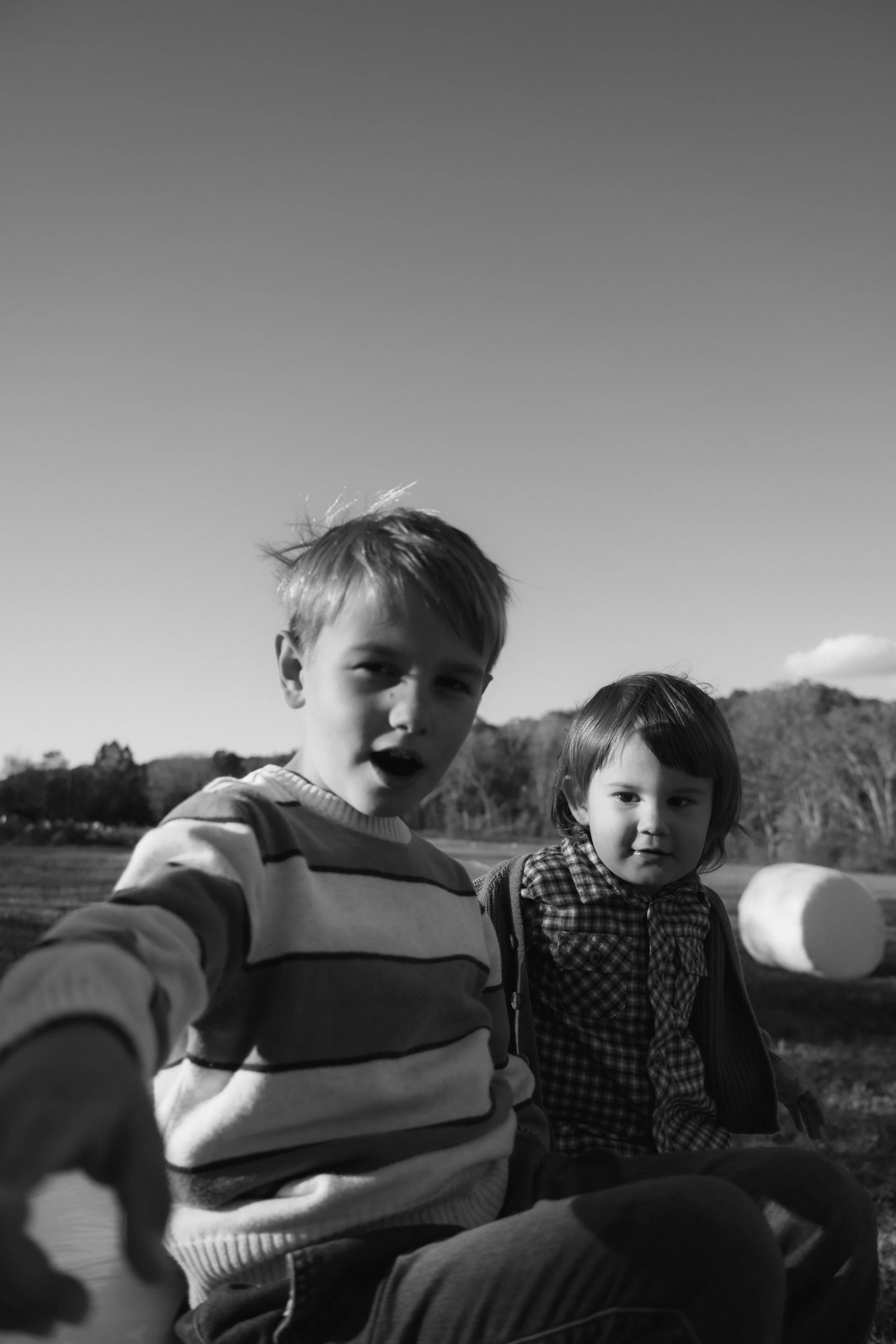 Two children sitting outdoors in a field with hay bales in the background, black and white photo
