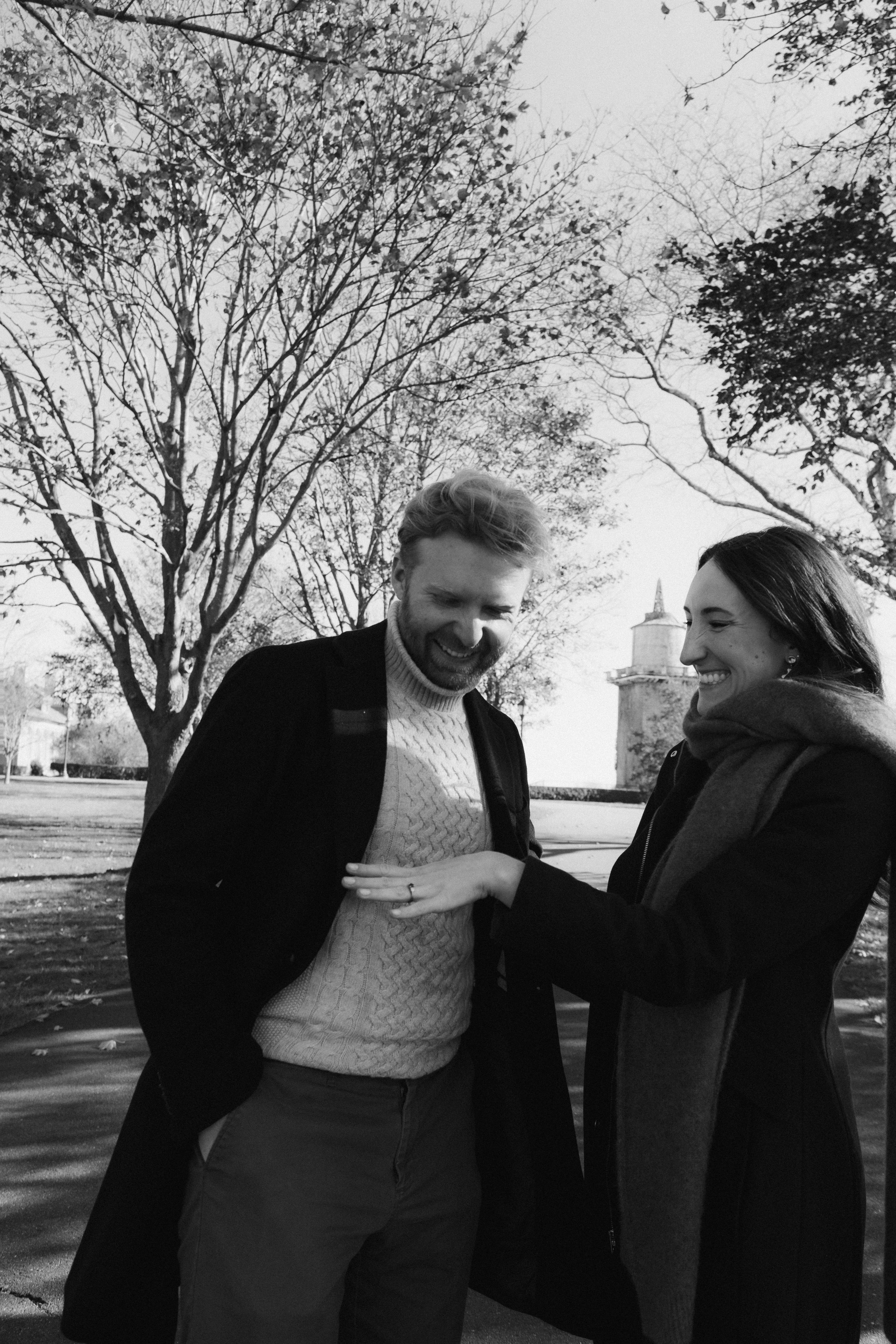 Black and white photo of a man and woman smiling and laughing outdoors, with trees and a building in the background, during daytime.