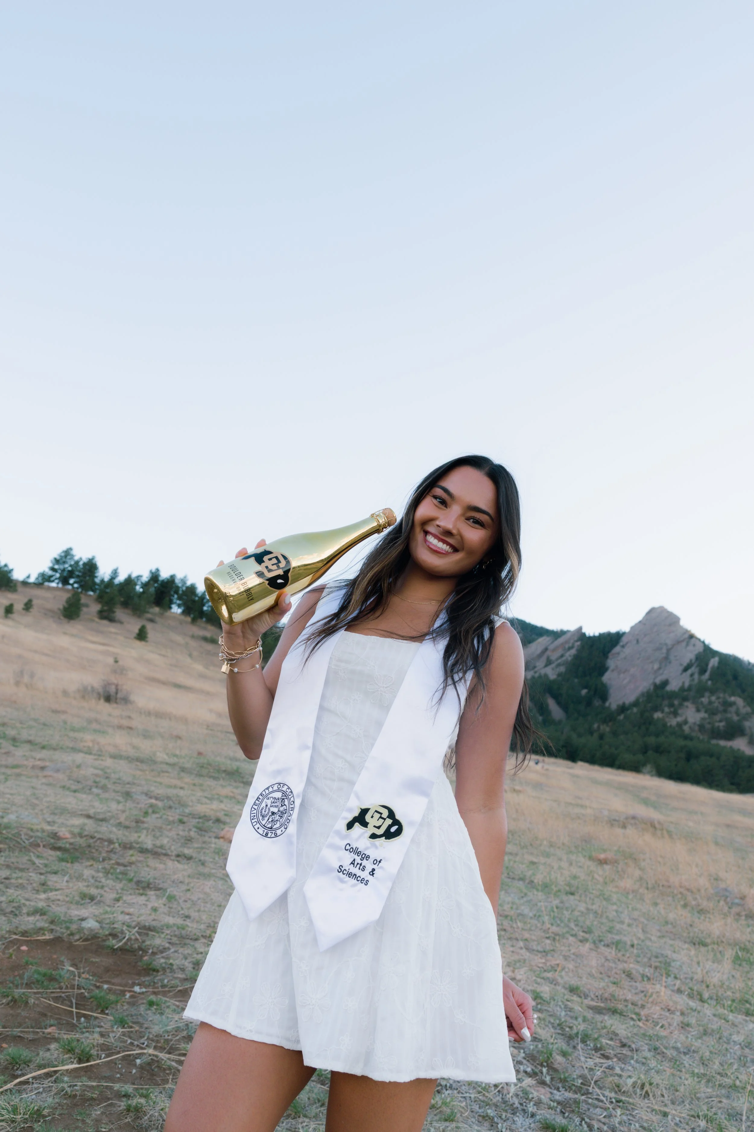 Young woman smiling outdoors holding a champagne bottle wearing a white dress and college graduation stole with mountains in the background.